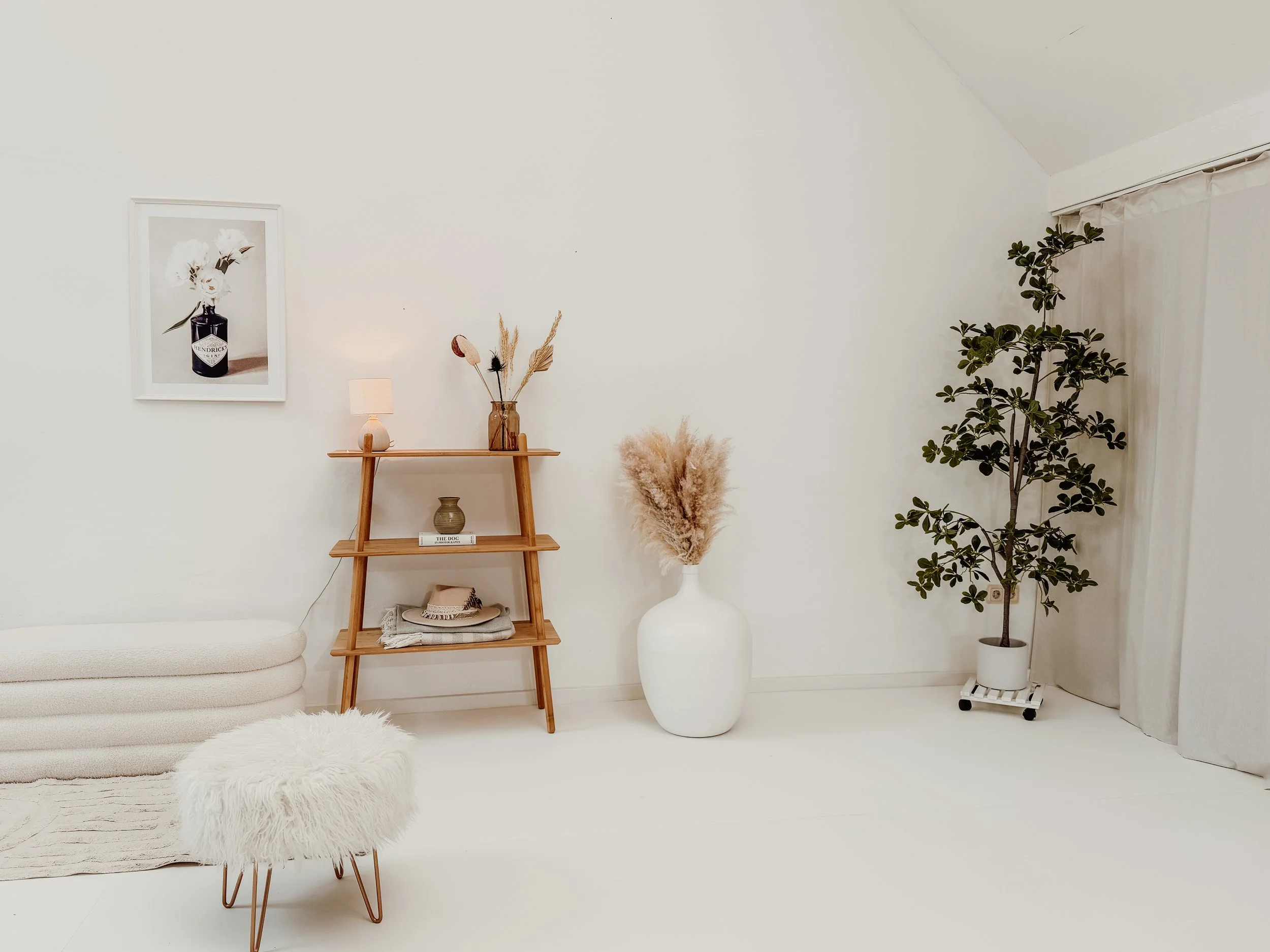 Ein minimalistisch eingerichtetes Fotostudio mit weißen Wänden, einem flauschigen Sitzhocker, einem Holzregal mit Dekorationen, einer großen weißen Vase mit Trockenblumen, und einer Topfpflanze vor weißen Vorhängen.