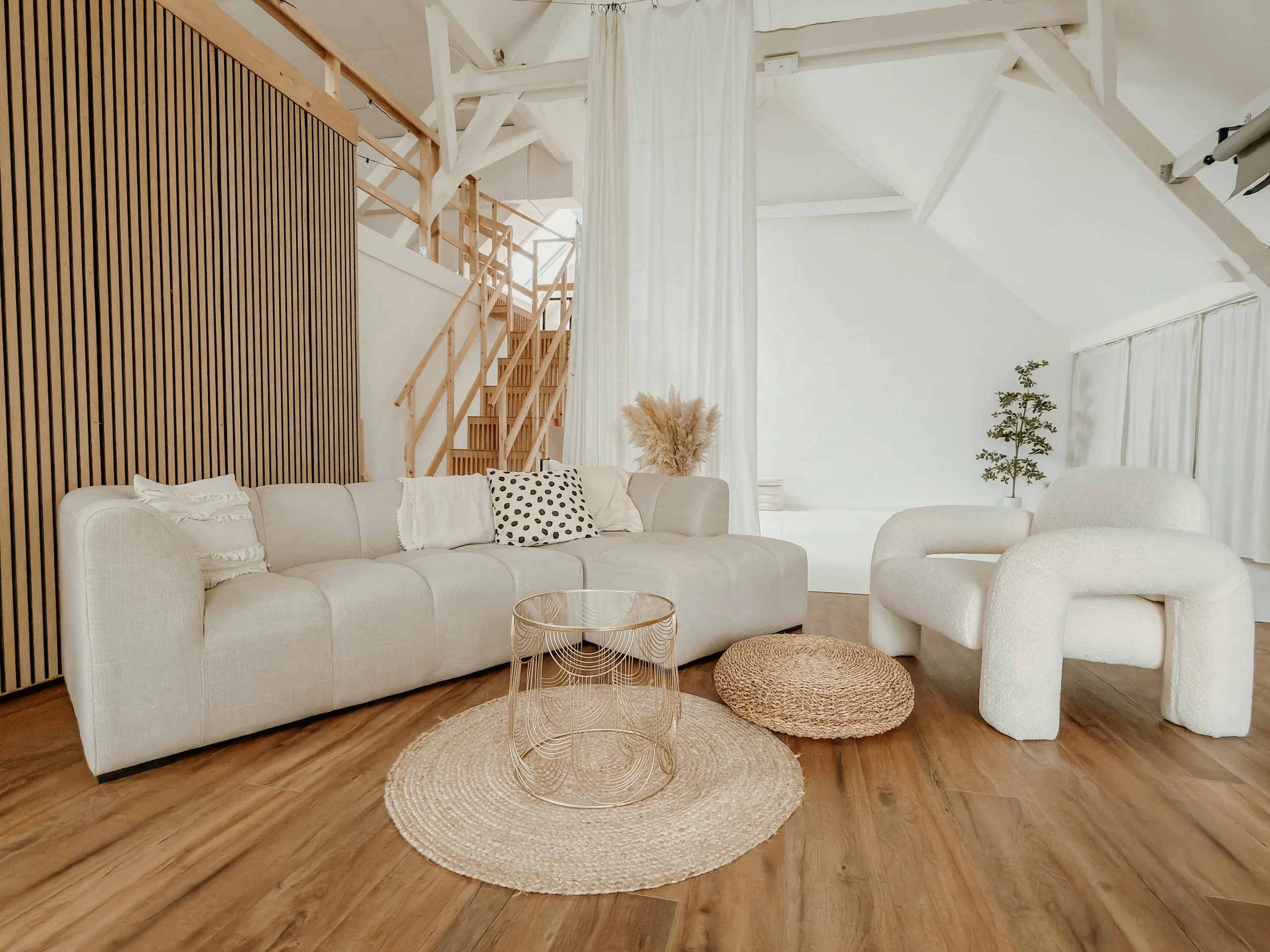 Gemütliches Fotostudio mit weißem Sofa, buntem Kissen, runden Rattan-Pouf und Teppichen, Holzstufen, Pflanzen und weißer Dekoration.