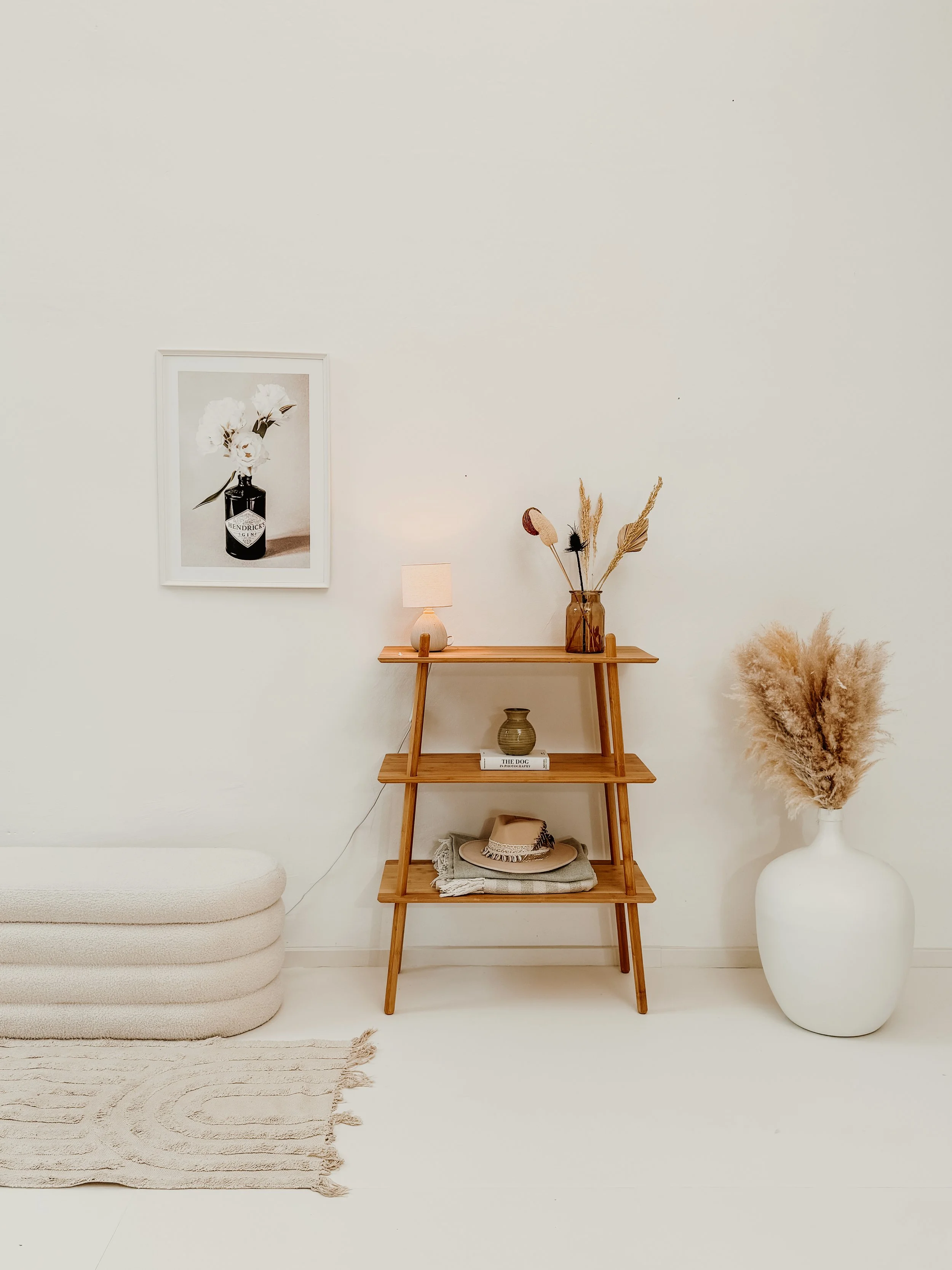 Minimalistische Inneneinrichtung mit einem weißen Wandhintergrund, einen weißen Bezugsofa, einen beige Teppich, ein Holzregal mit Dekorationen, einer kleinen Lampe, einem Buch, einem Hut und getrockneten Pflanzen in einer großen weißen Vase.