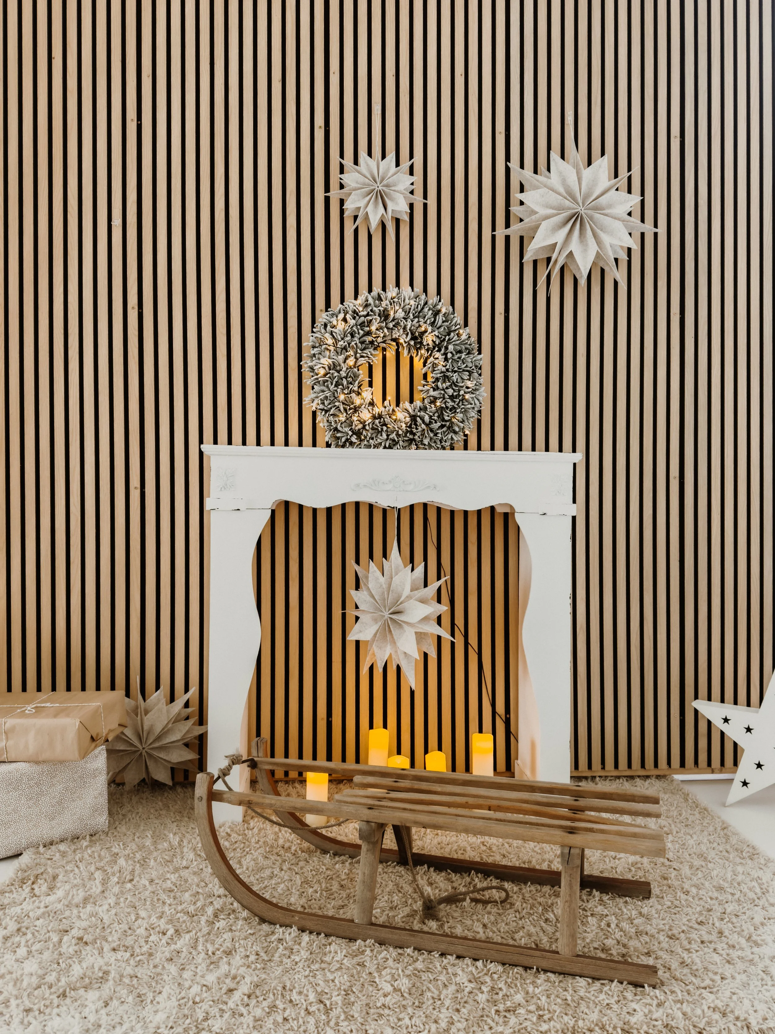 Weihnachtliche Dekoration mit Papiersternen, Lichterkranz, Kerzen und Holzschlitten vor einem Holzpaneel-Hintergrund.