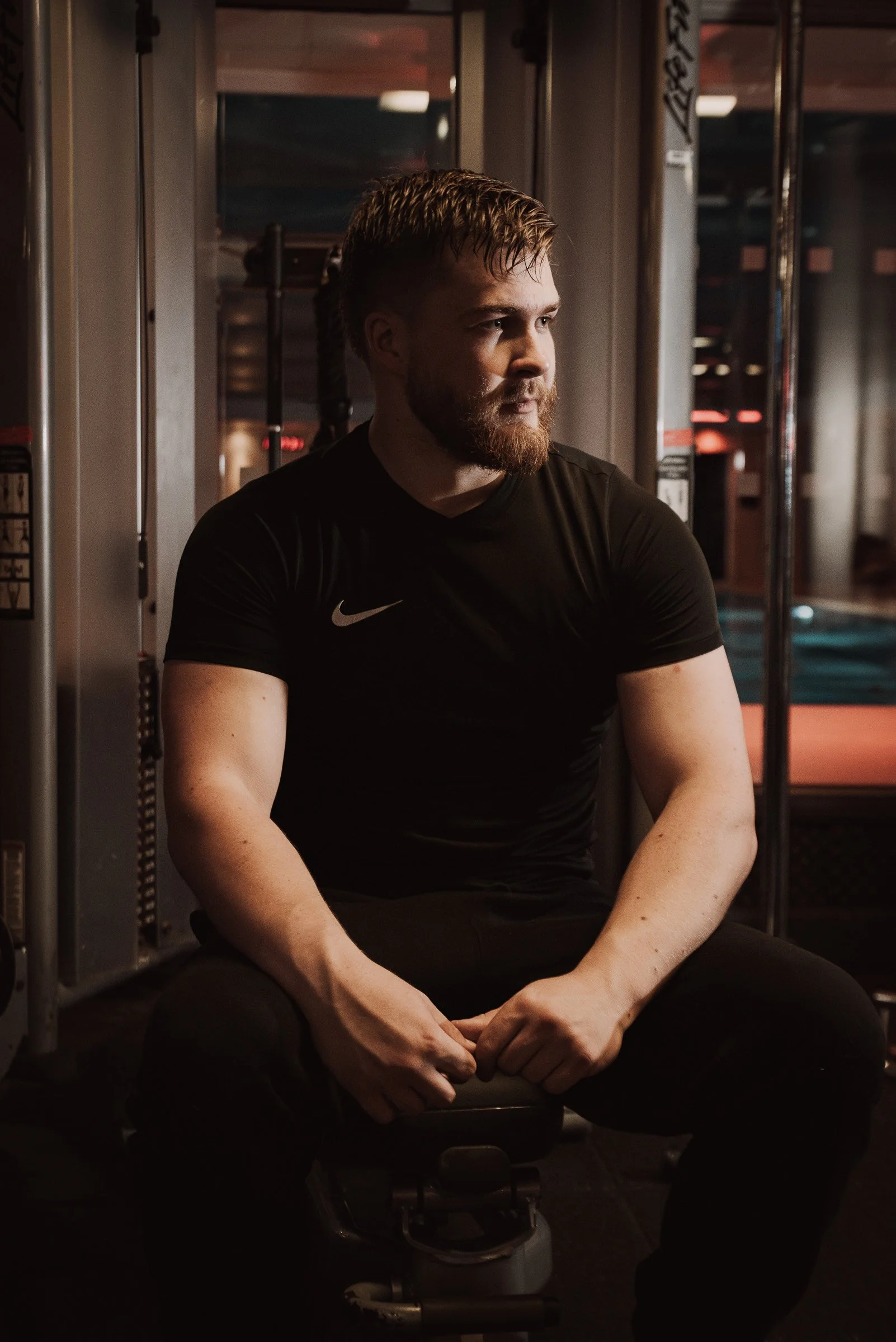 A man with a beard and wet hair sitting on a gym bench, wearing a black Nike t-shirt, in a gym setting with exercise equipment in the background.