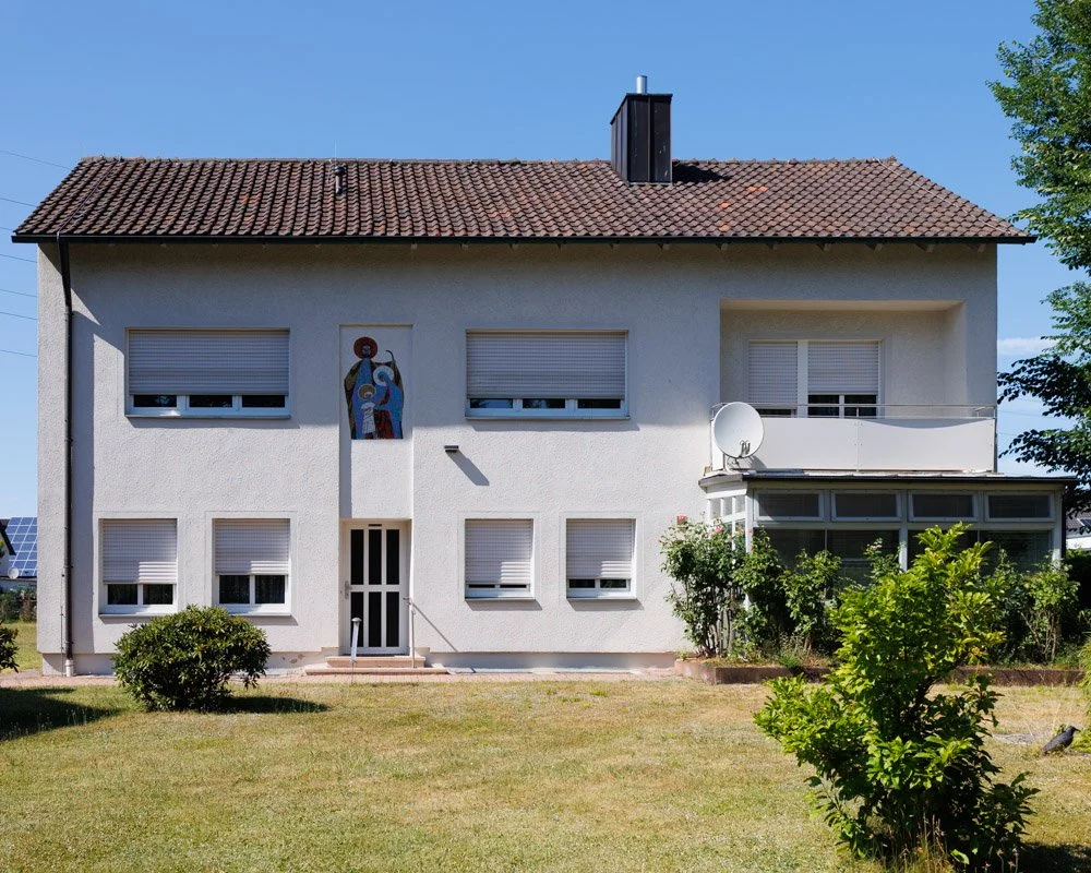 Ein weißes zweistöckiges Haus mit rotem Ziegeldach, mehreren Fenstern mit Rollläden, einem Eingang mit Treppe, einer Satellitenschüssel, einem bemalten Wandbild, Garten mit Büschen und einem Baum, im Hintergrund blauer Himmel.