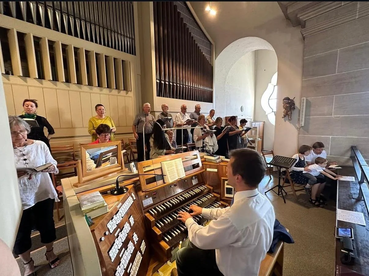 Chor in einem Kirchenraum, begleitet von einem Organisten und einem kleinen Kinderchor, während sie proben.