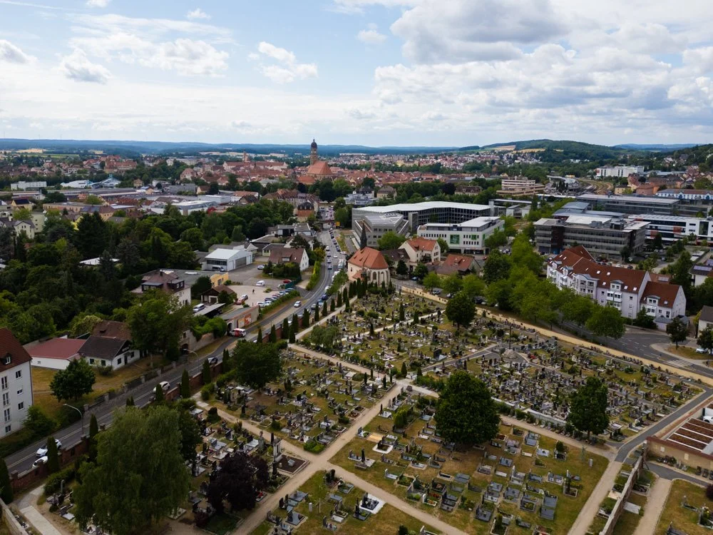Luftaufnahme einer Stadt mit Friedhof im Vordergrund und Wohnhäusern sowie Hochhäusern im Hintergrund, bei bewölktem Himmel.