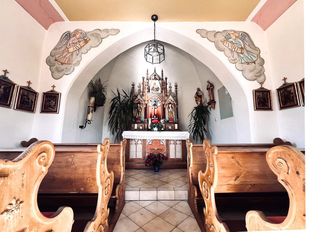 Innenraum einer kleinen katholischen Kirche mit Holzkapellen und einem Altar, der mit Blumen geschmückt ist. An den Wänden sind Heiligenbilder und zwei gemalte Engel, die Wolken tragen, zu sehen.