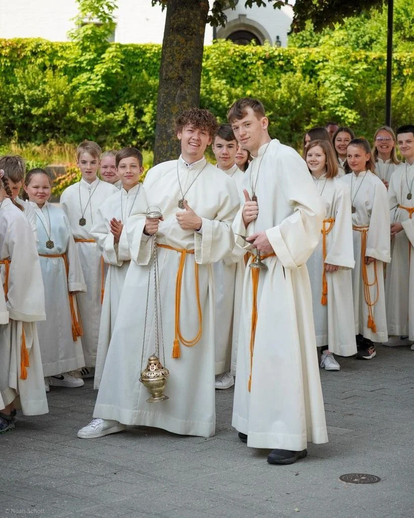 Eine Person, wahrscheinlich bei einer religiösen Zeremonie, hält die Hände einer anderen Person in Gebetshaltung. Im Hintergrund sind Blumen sichtbar.