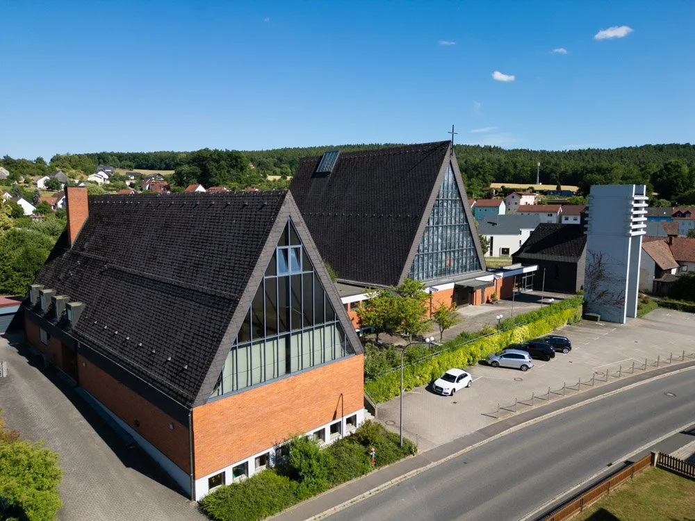 Luftaufnahme einer modernen Kirche mit großen Glasfenstern und einem steilen Dach, umgeben von einem Parkplatz und grünem Garten, in einer ländlichen Umgebung.
