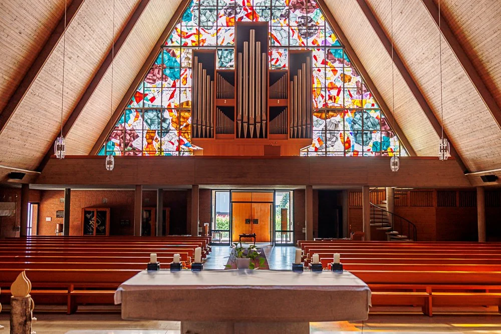 Innenansicht einer Kirche mit Holzarchitektur, großen farbigen Glasfenstern und einer Orgel im Altarbereich.