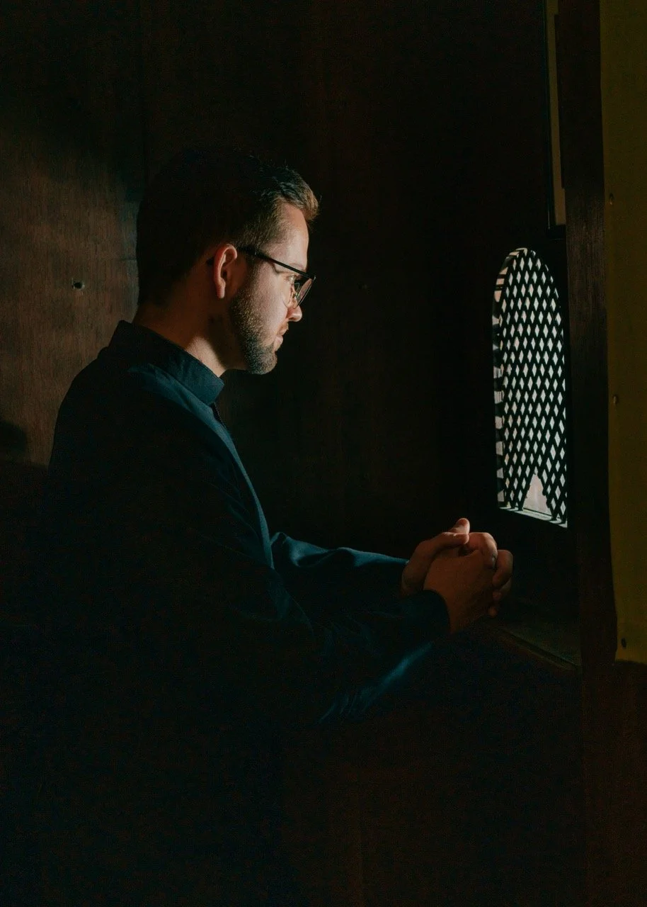 Ein Mann in dunkler Kleidung und Brille sitzt betend vor einem kleinen Fenster mit Gitter in einer dunklen Umgebung.