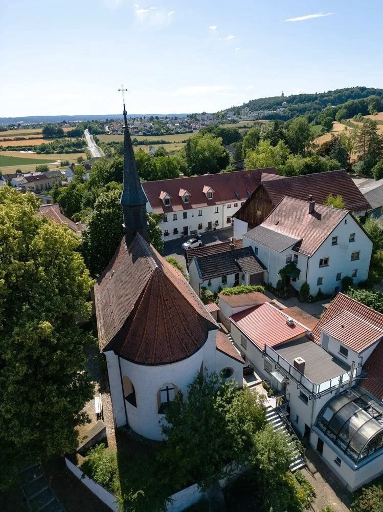 Luftaufnahme einer kleinen Kirche mit Spitzdach und Kirchturm in einer ländlichen Gegend mit mehreren Häusern und grünen Bäumen.