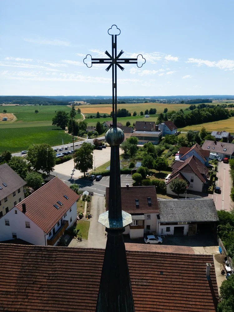 Blick auf eine weite ländliche Szenerie mit Häusern, Feldern und Wäldern, aufgenommen von einer Kirche mit einer markanten Eisenkreuz-Spitze im Vordergrund.