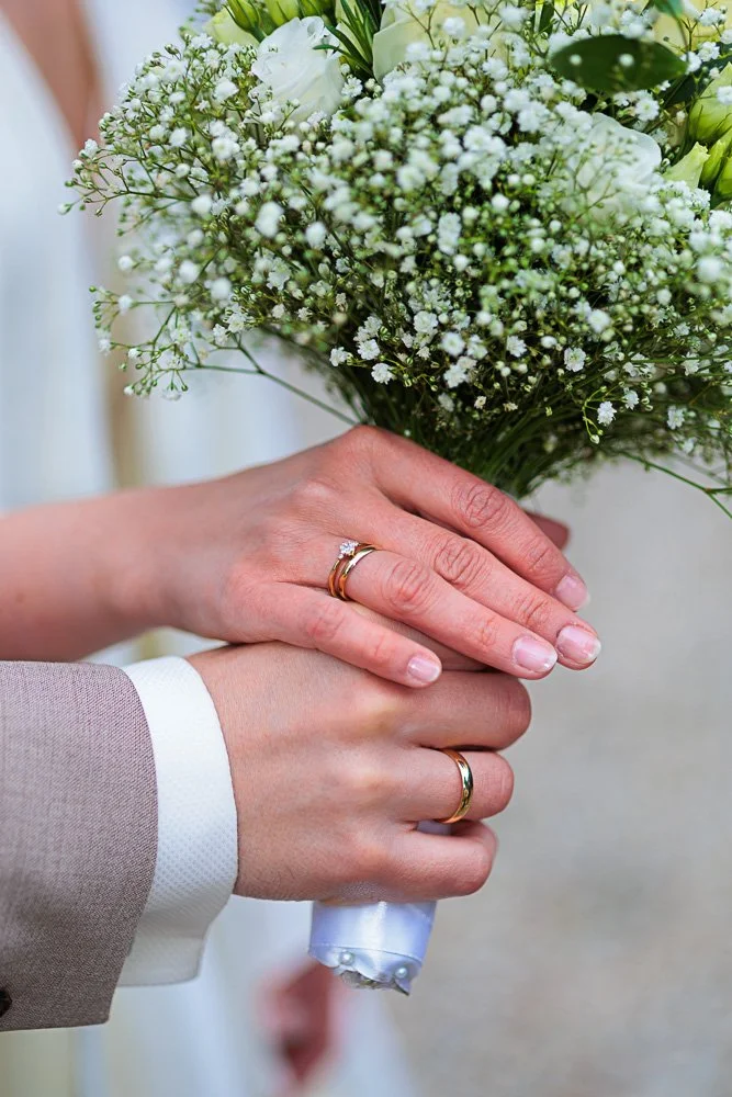 Hände eines Paares mit Eheringen, das einen Blumenstrauß aus weißen Blumen hält.