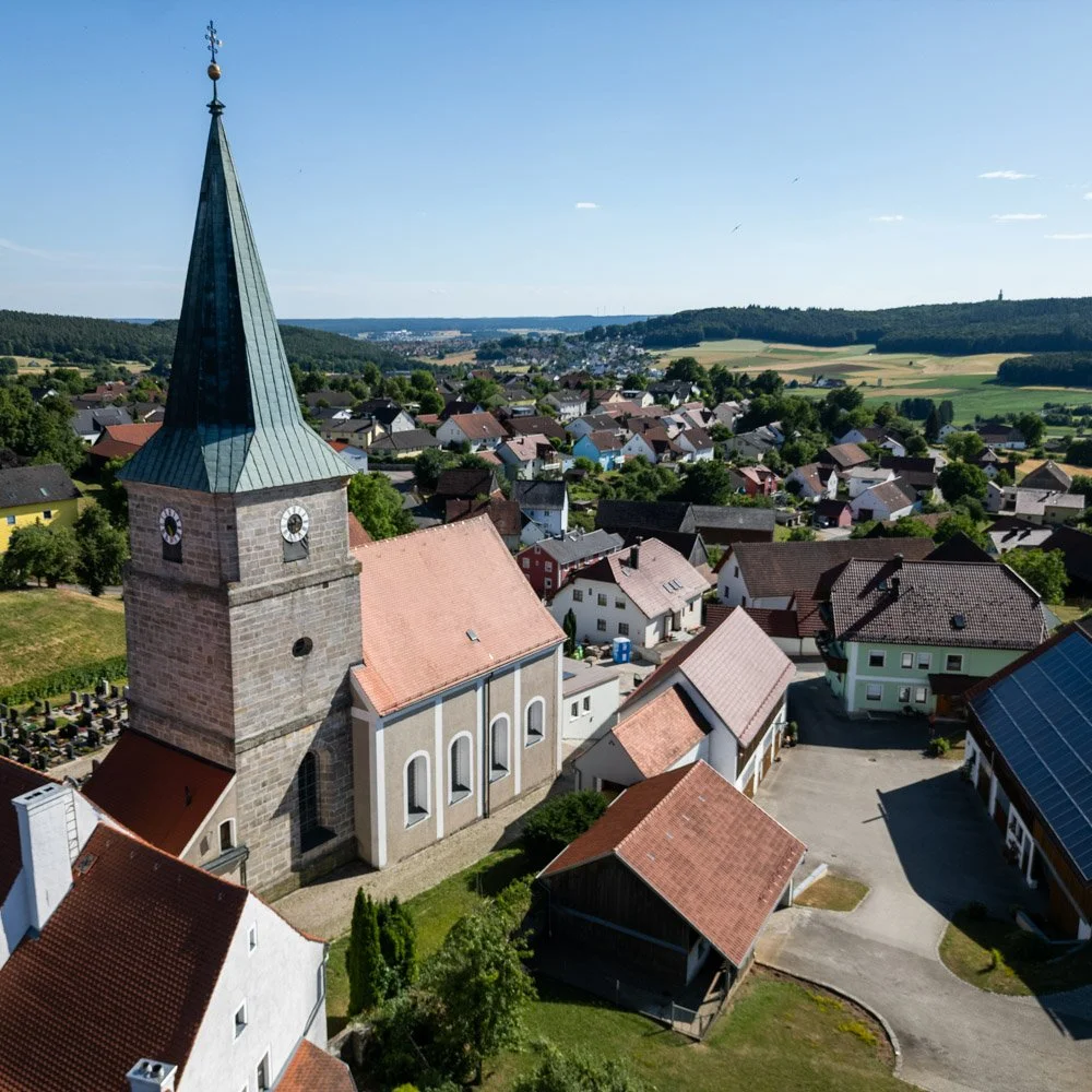 Ein Dorf mit einer Kirche mit einem hohen spitzen Turm und umliegenden Häusern in einer ländlichen Gegend bei sonnigem Wetter.
