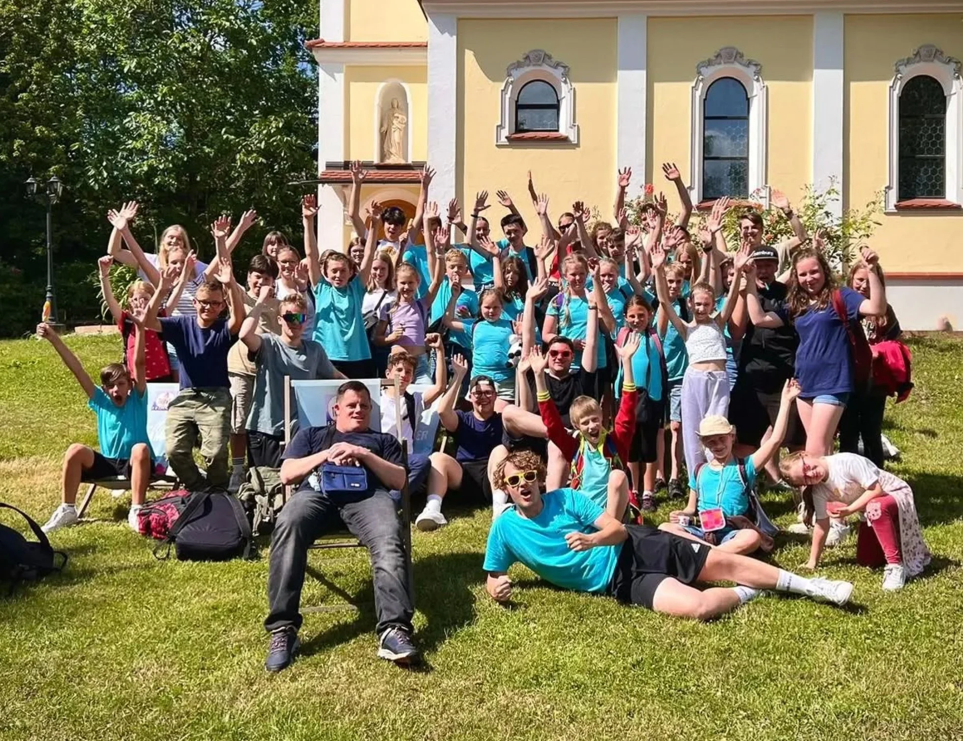 Gruppe von Kindern und Jugendlichen vor einem gelben Gebäude, alle lachen und winken in die Kamera, einige sitzen auf dem Rasen, andere stehen, sonniger Tag.
