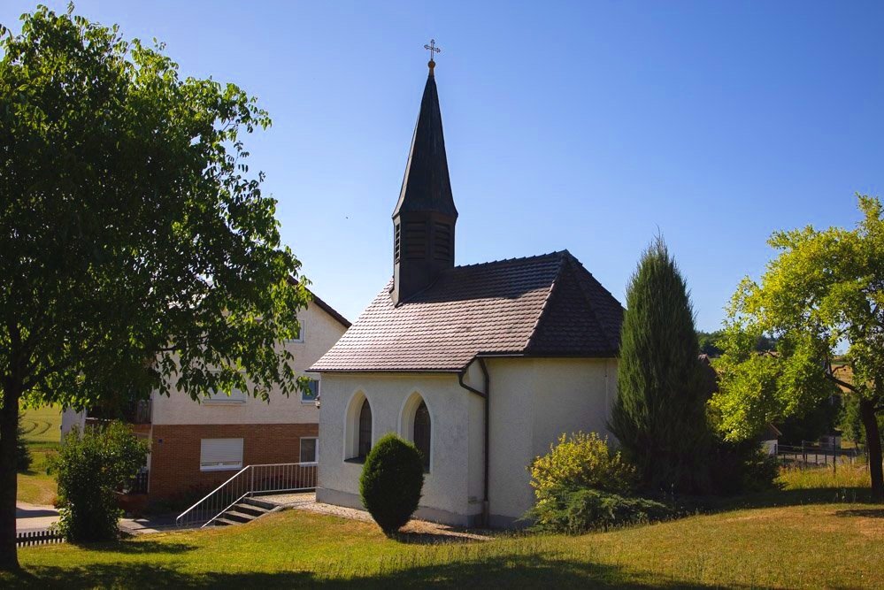 Kleine Kirche mit steinähnlicher Fassade, schmalem steinernen Turm und Kreuz oben auf dem Dach, umgeben von Bäumen und grünem Rasen, bei sonnigem Himmel.