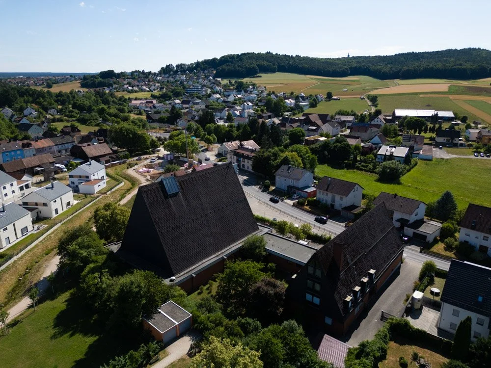 Luftaufnahme eines kleinen Dorfes mit Häusern, Straßen, Wäldern und landwirtschaftlichen Flächen in einer ländlichen Gegend.