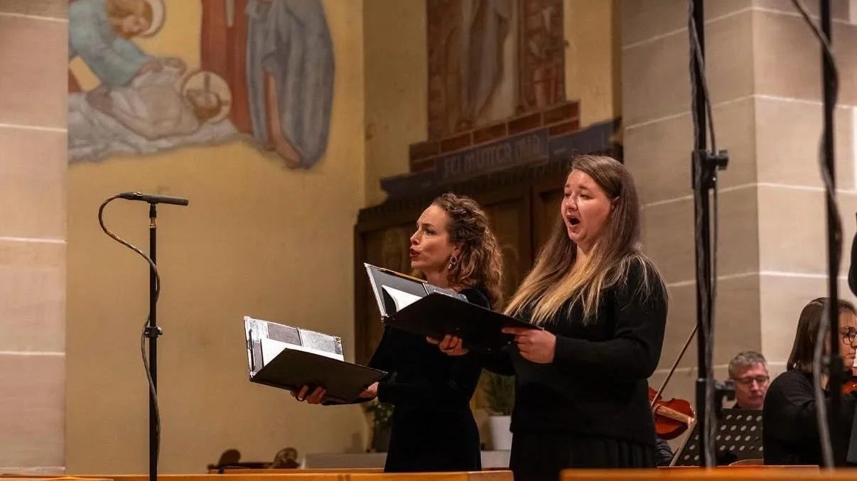 Zwei weibliche Sängerinnen in einem Konzertsaal, die beim Singen eine Kantate oder Oper singen, während im Hintergrund Musiker mit Instrumenten sitzen.
