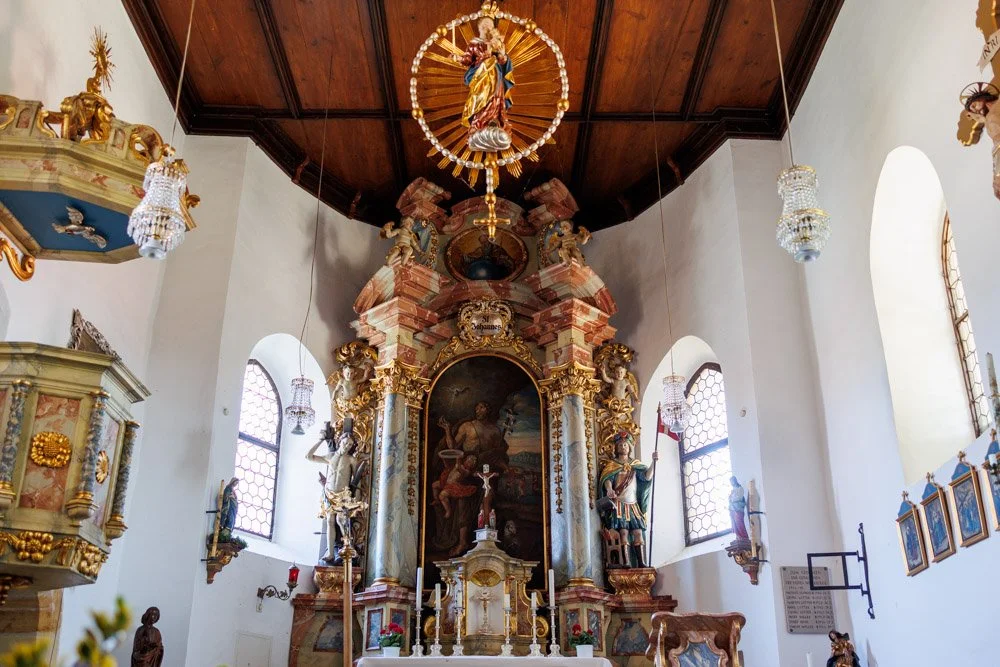 Innenraum einer barocken Kirche mit Altar, barocken Skulpturen, bunten Fenstern, Kronleuchtern und einer geschnitzten Holzdecke.