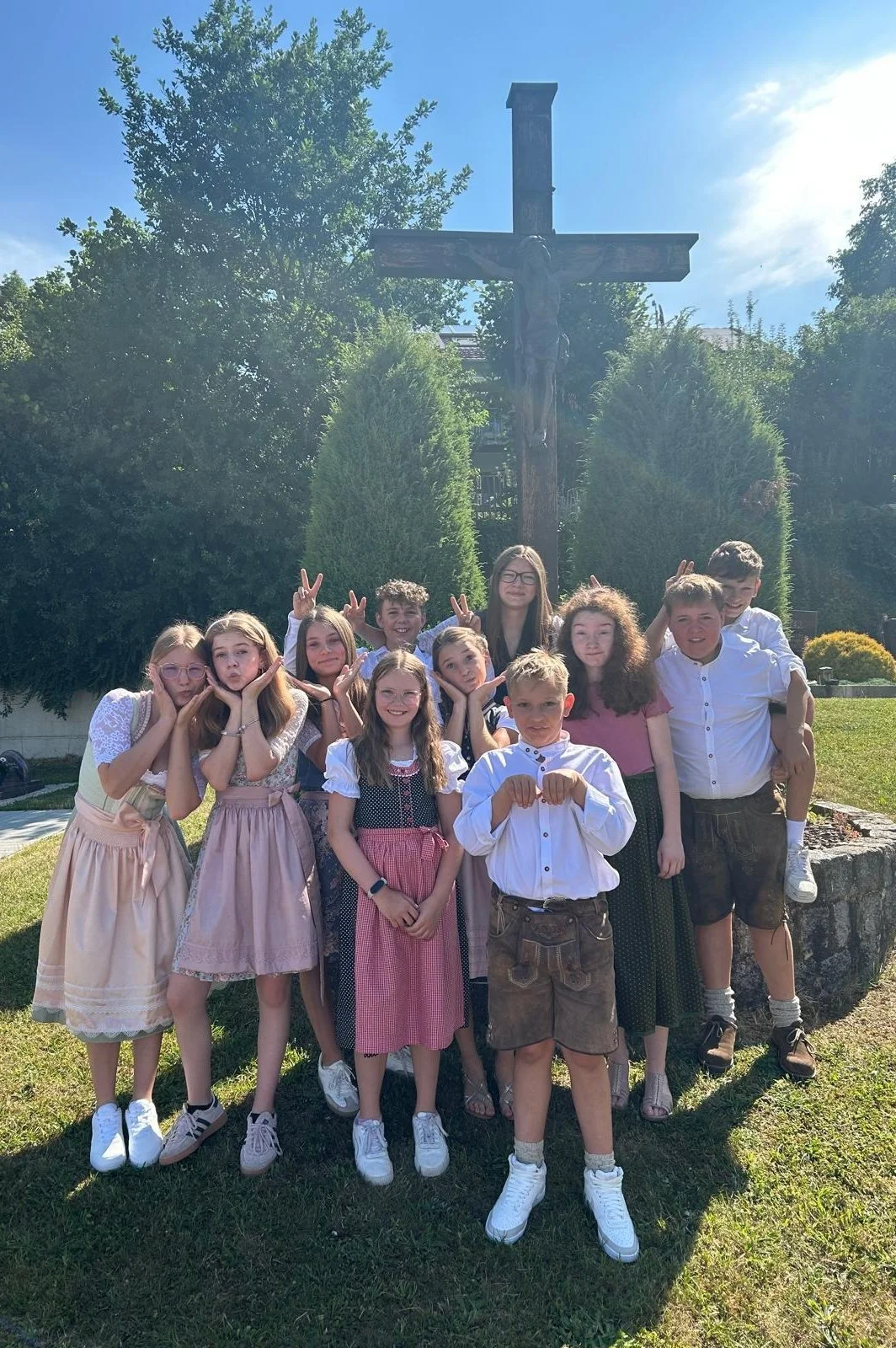 Eine Gruppe von Kindern in traditionellen Trachten posiert vor einem großen Kreuz im Freien bei sonnigem Wetter.