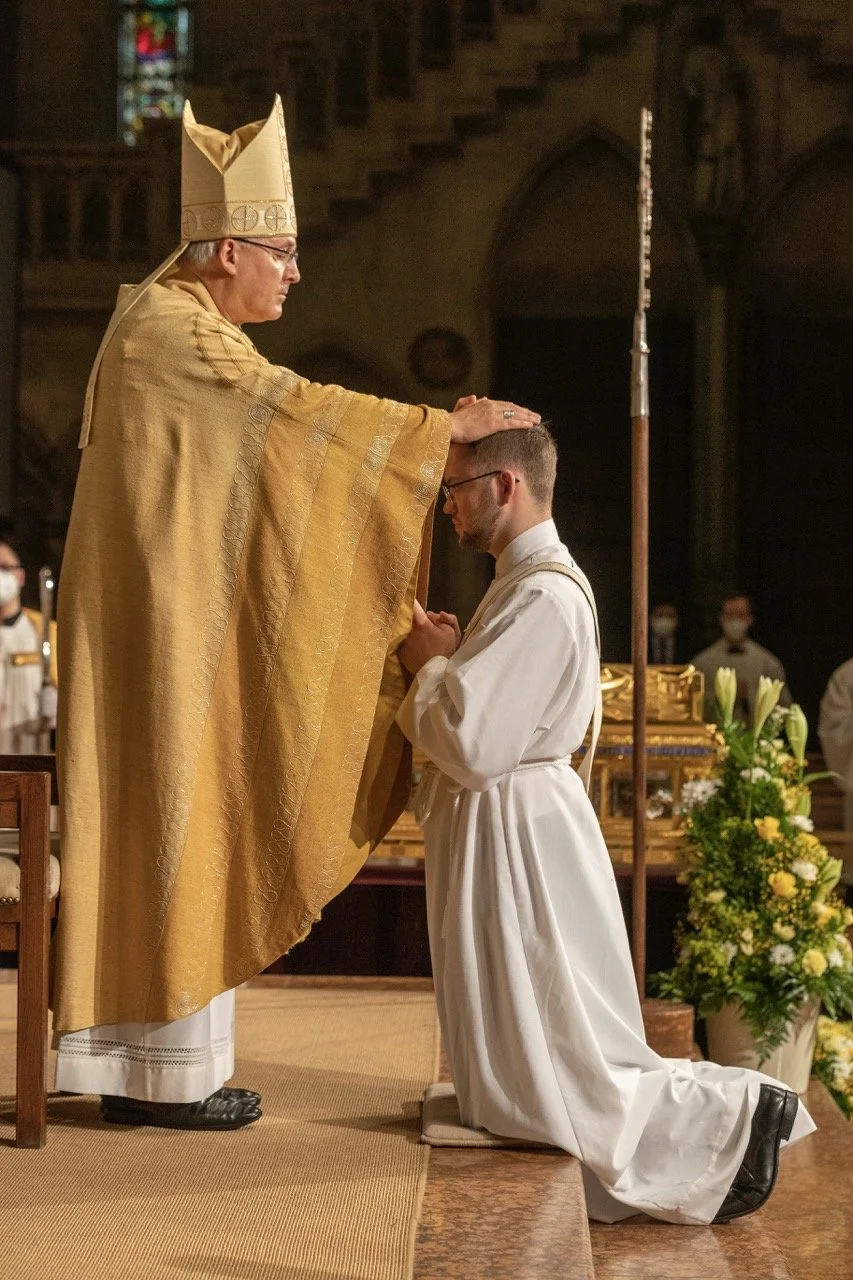 Ein Priester in goldener Kleidung segnet einen jungen Mann, der in weißem Gewand knieend mit gefalteten Händen betet, in einer Kirche mit Blumen und anderen Personen im Hintergrund.
