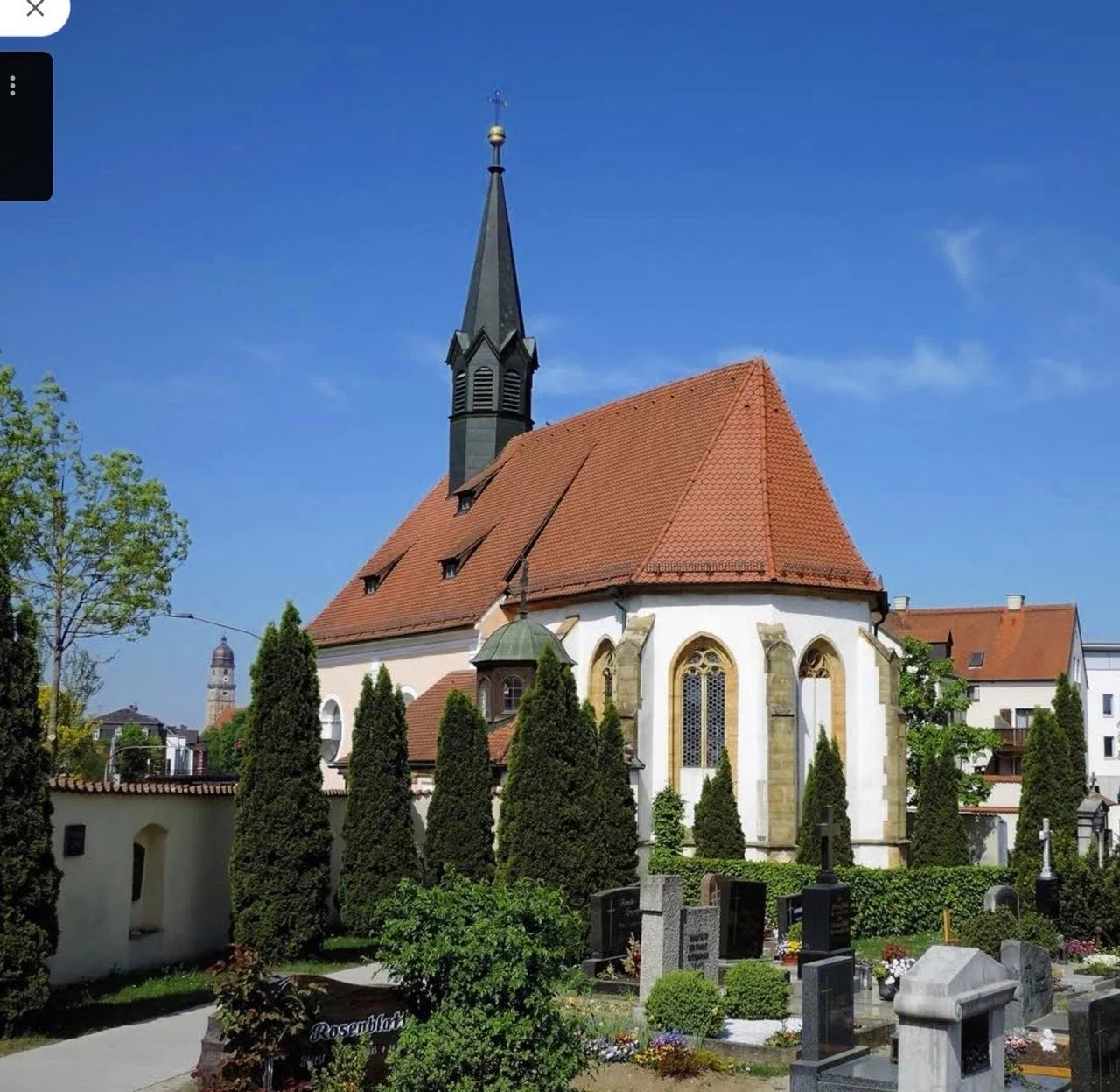 Eine alte Kirche mit roten Dachte und einem hohen Kirchturm, umgeben von einem Friedhof mit Bäumen und Grabsteinen, bei sonnigem Himmel.