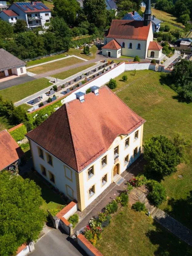 Luftaufnahme eines Gebäudes mit rotem Dach und kleinen Gärten, umgeben von Bäumen und einer Kirche im Hintergrund.