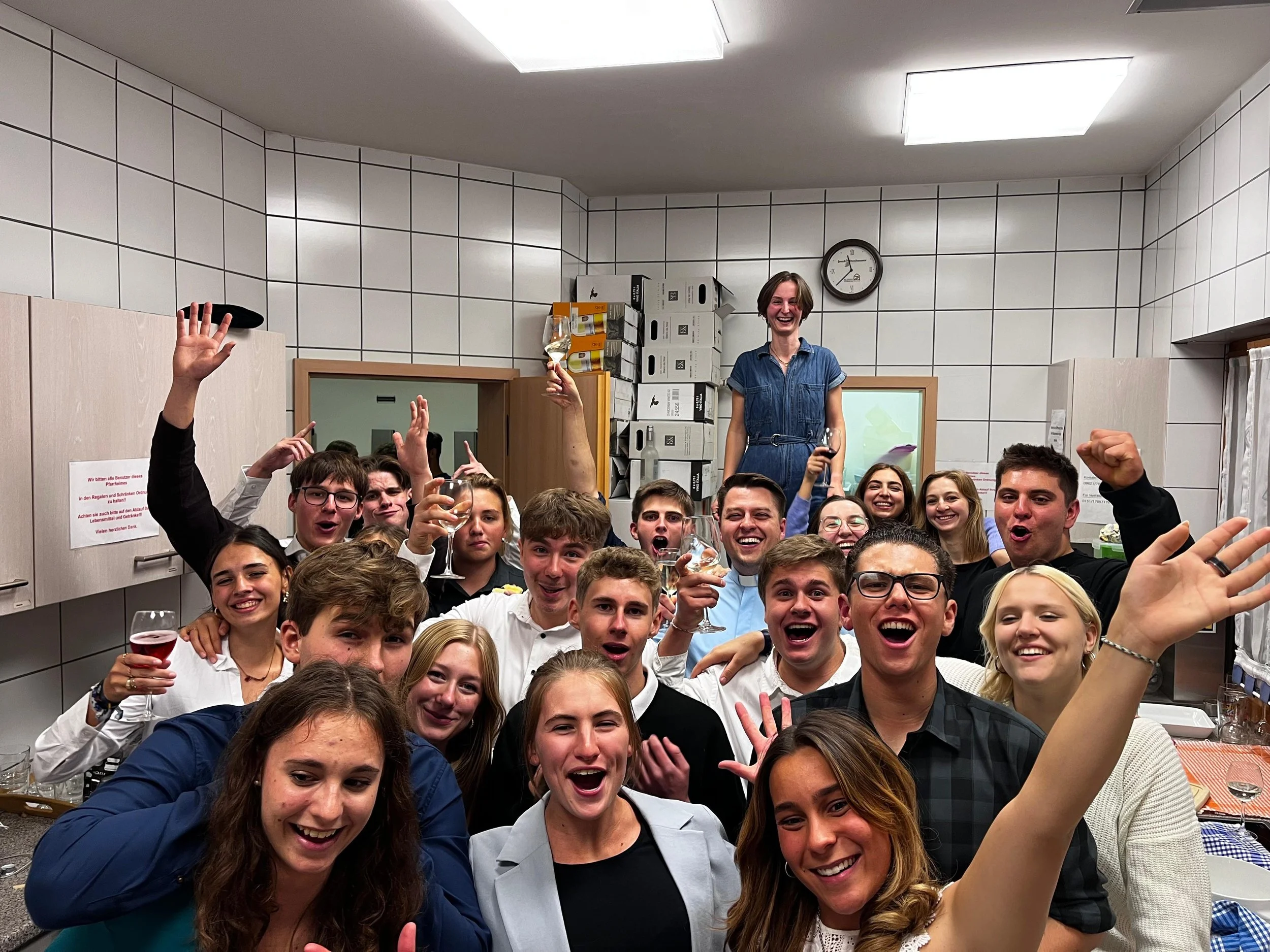 Menschen feiern in einer Küche, einige halten Weingläser, andere haben die Faust hoch. Eine Frau steht auf einem Tisch, alle lachen und freuen sich.