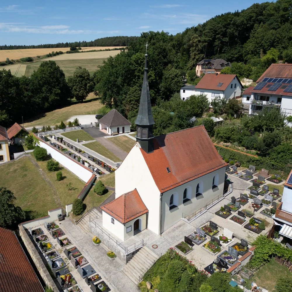 Luftaufnahme einer kleinen Kirche mit Friedhof im Vordergrund, umgeben von Wohnhäusern und grünen Hügeln.