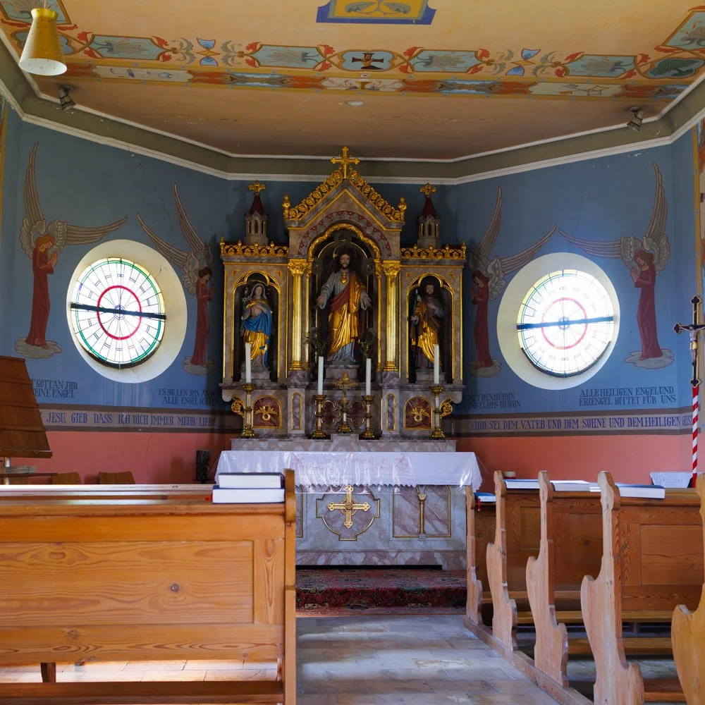 Innenansicht einer Kirche mit Altartafel, zwei bunten Kirchenfenstern und Holzbänken.