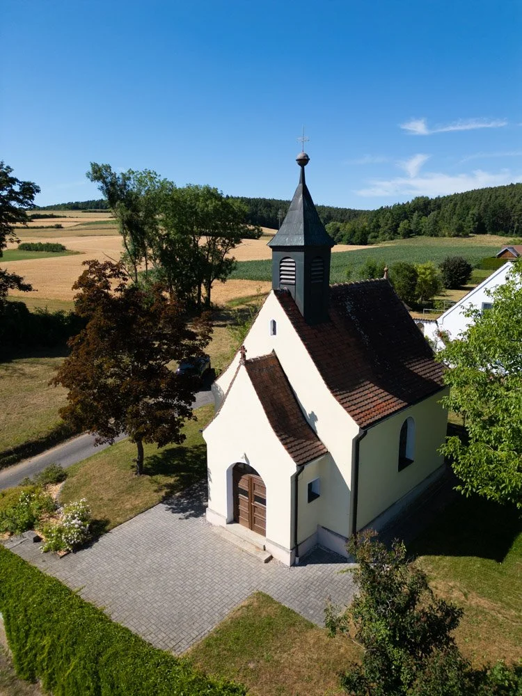 Kleine weiße Kirche mit dunklem Dach und schmalem Turm, umgeben von Bäumen und Feldern unter blauem Himmel.