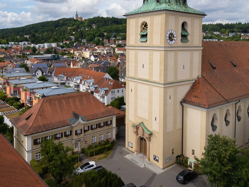 Blick auf eine Kirche mit einem hohen Turm und Zifferblättern, umgeben von Wohnhäusern in einer Stadt mit Hügel im Hintergrund.