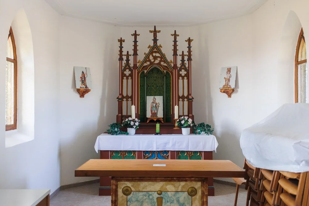 Kleiner Altar in einer Kirche mit Jesus-Ikone, Kerzen, Blumen und Bildern von Heiligen, umgeben von weißen Wänden und Fenstern.