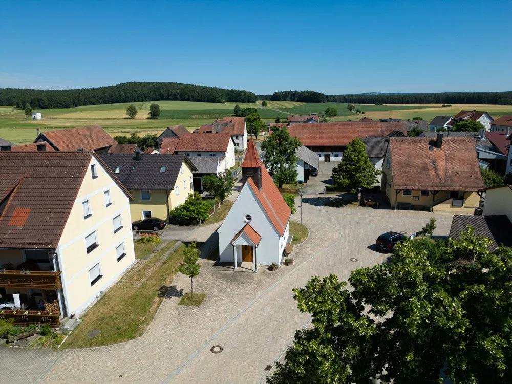 Ein ruhiges Dorf mit Häusern, umgeben von grünen Wiesen und Bäumen, bei sonnigem Himmel.