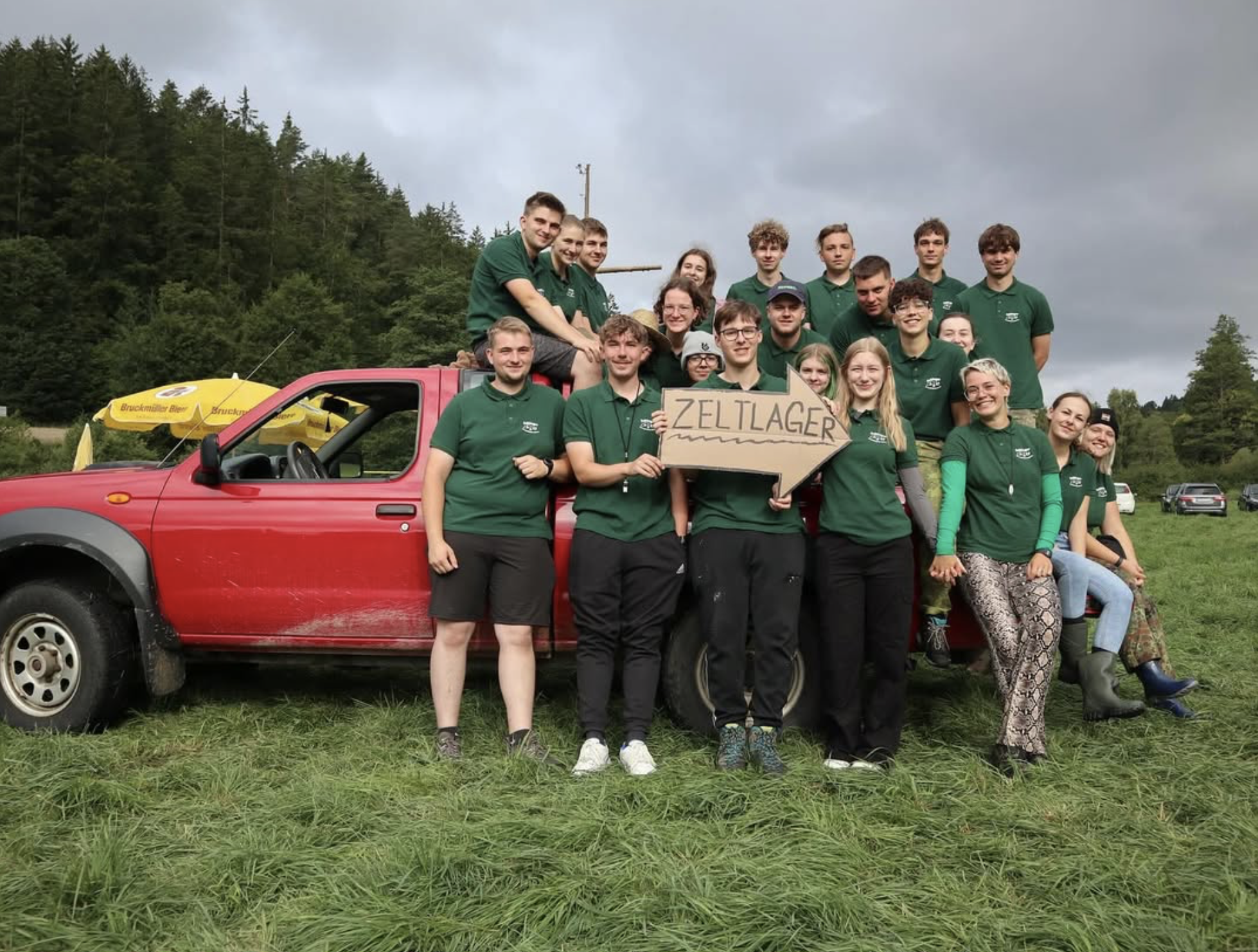 Gruppenfoto von Jugendlichen mit grünen T-Shirts auf einem Hänger in einem grünen Feld, eine Person hält ein Schild mit der Aufschrift 'Zeltlager', im Hintergrund Bäume und Wolken.