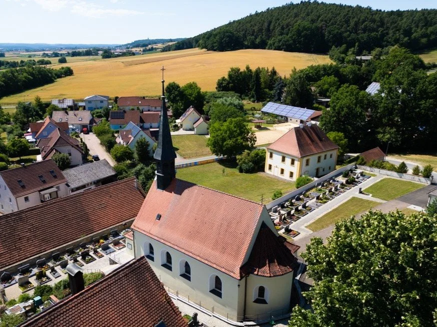 Luftaufnahme eines kleinen Dorfs mit Kirche, umliegenden Häusern, Feldern und Wäldern in der Landschaft.