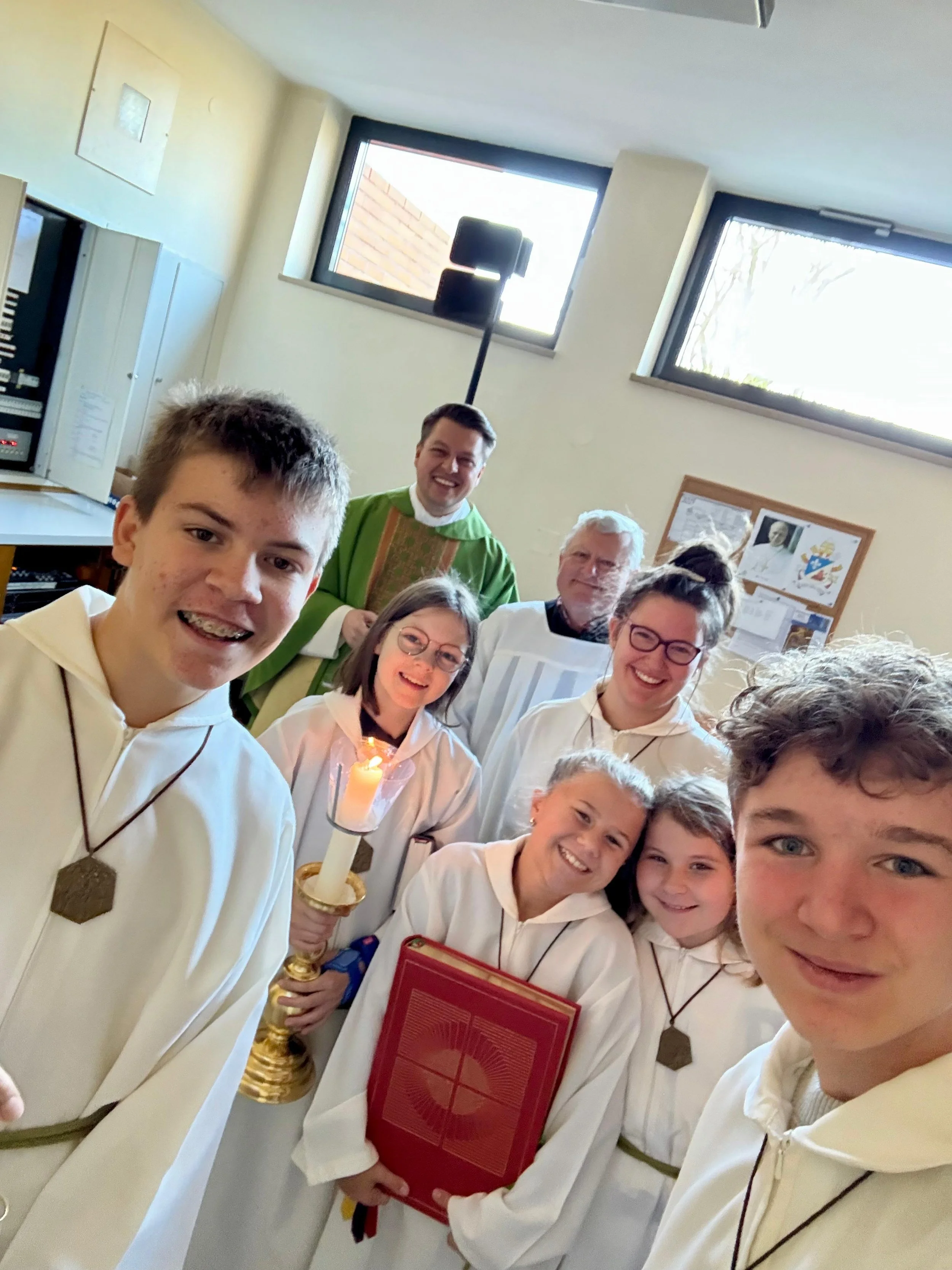 Gruppen von Menschen, viele Kinder und einige Erwachsene, feiern zusammen, einige tragen weiße Gewänder mit Medaillen, ein Kind hält eine Kerze und ein anderes einen roten Gegenstand, im Raum mit Fenstern und einer Pinnwand.