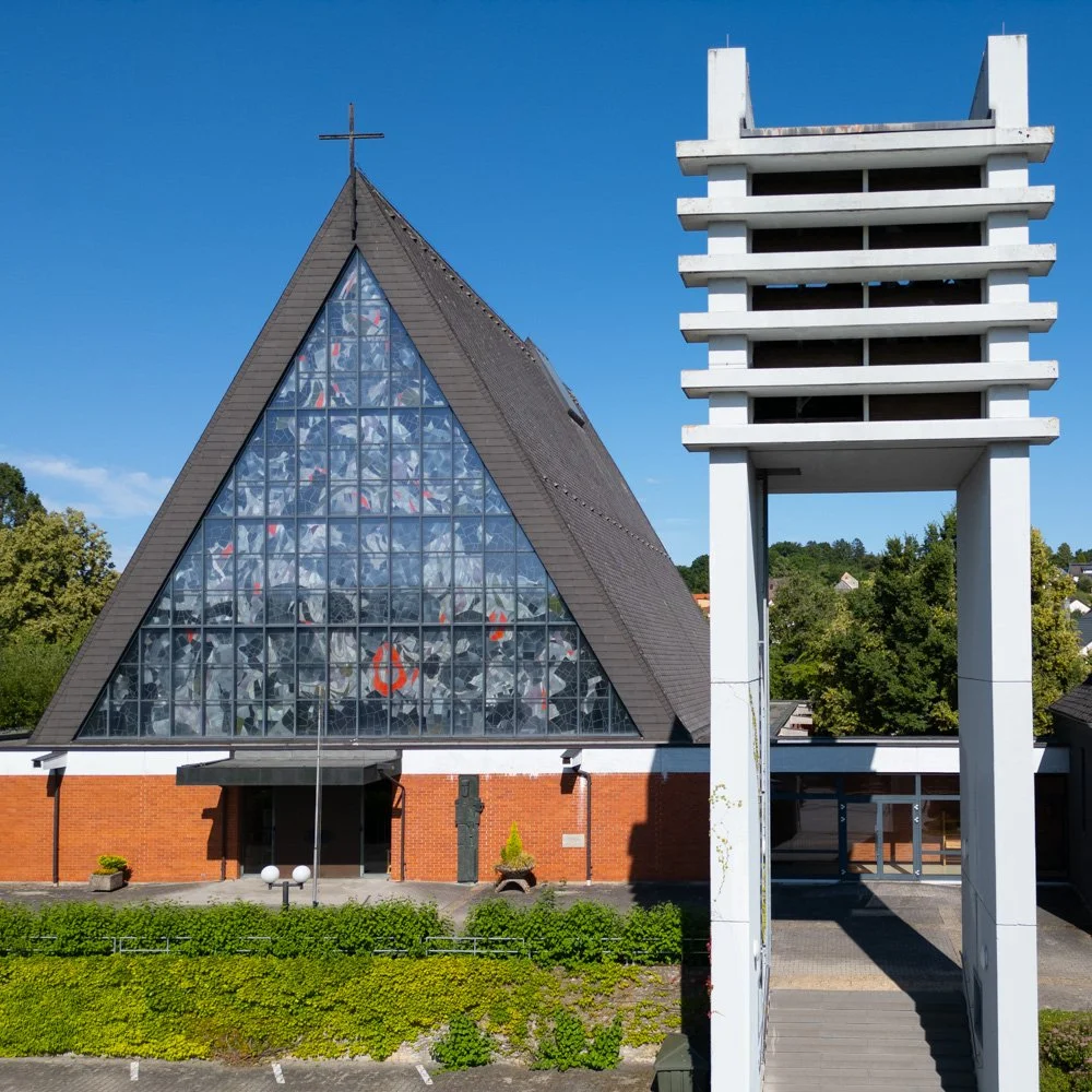 Eine moderne Kirche mit einem großen, glasüberdachten, dreieckigen Kirchenschiff und einem hohen Glockenturm vor einem blauen Himmel.