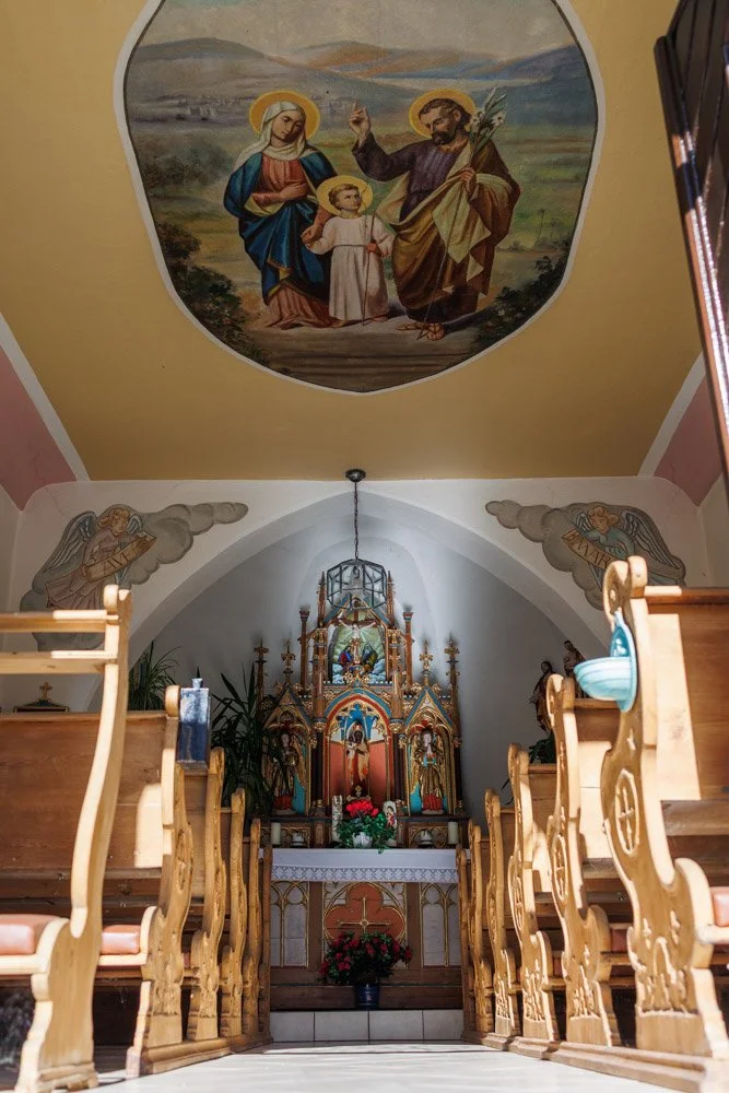 Innenraum einer Kirche mit Holzbankreihen, einem Altar mit religiösen Figuren und Kerzen, sowie biaflächigen Decken- und Wandmalereien, darunter eine Szene mit heiligen Figuren.