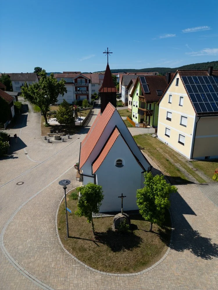 Vereinzelte moderne Häuser mit Solardächern, kleine Kirche mit Kirchturm und Kreuz, umgeben von Bäumen und einem gepflasterten Platz an einem sonnigen Tag.