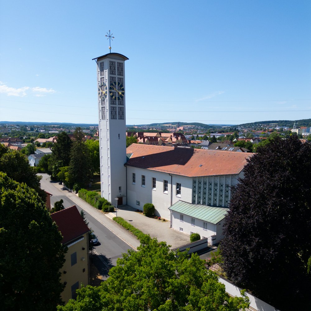 Ein weißer Kirchturm mit einem Uhr und einer Wetterfahne auf dem Dach, umgeben von Bäumen und Häusern in einer Stadt bei sonnigem Himmel.