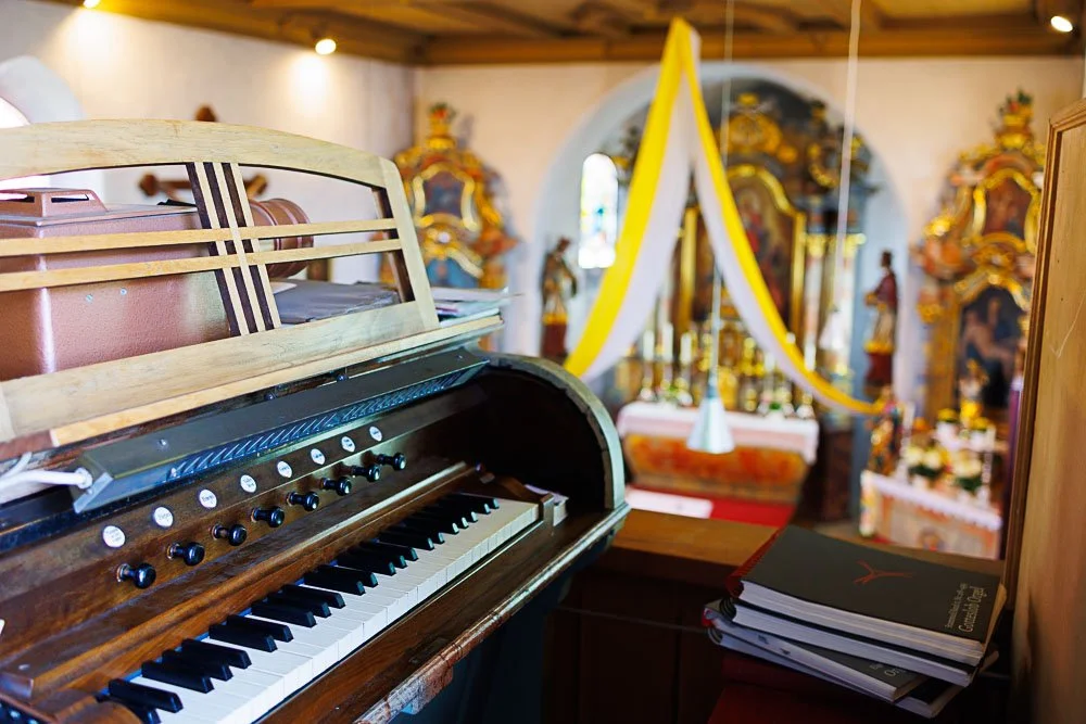 Ein alter, offener Elektroklavier mit dunkler Holzoptik im Vordergrund, im Hintergrund eine festlich geschmückte Kirche mit bunten Holzschnitzereien und einer weißen und gelben Dekoration