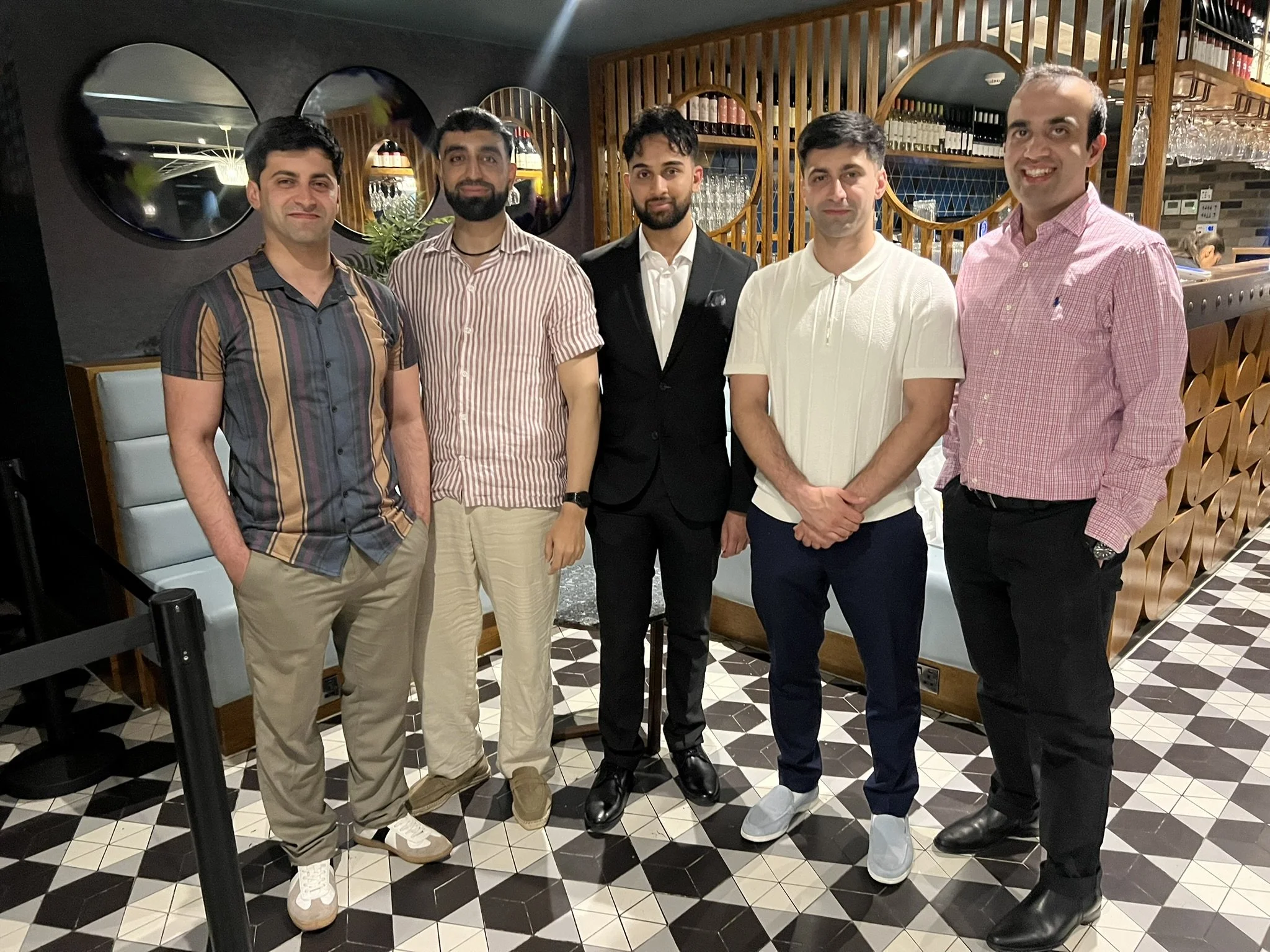 Group of six men standing in a restaurant, posing for a photo. They are dressed in casual and semi-formal attire, with a bar and decorative mirrors in the background.