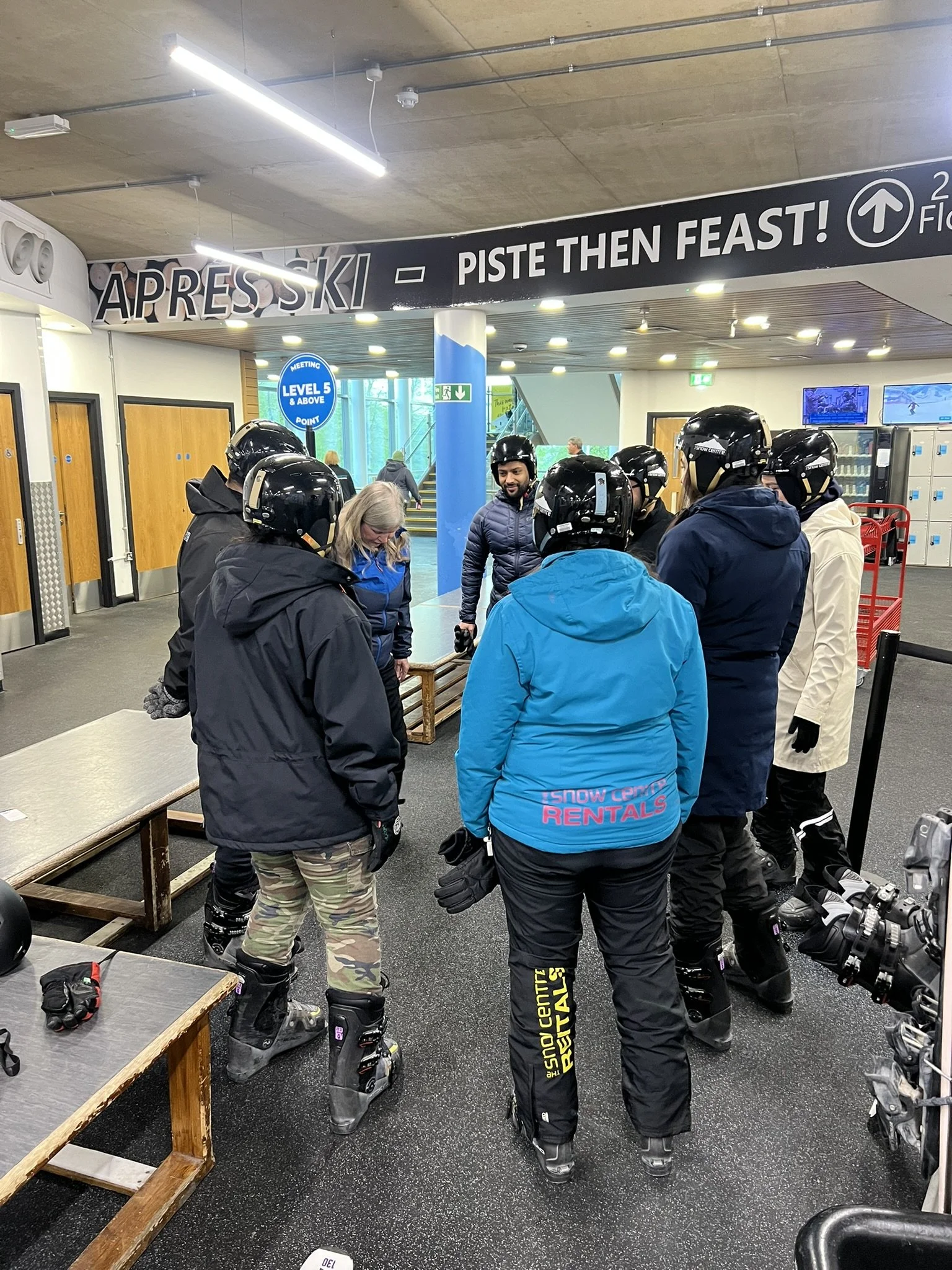 A group of people in ski gear gathered around a ski instructor at an indoor ski rental shop.