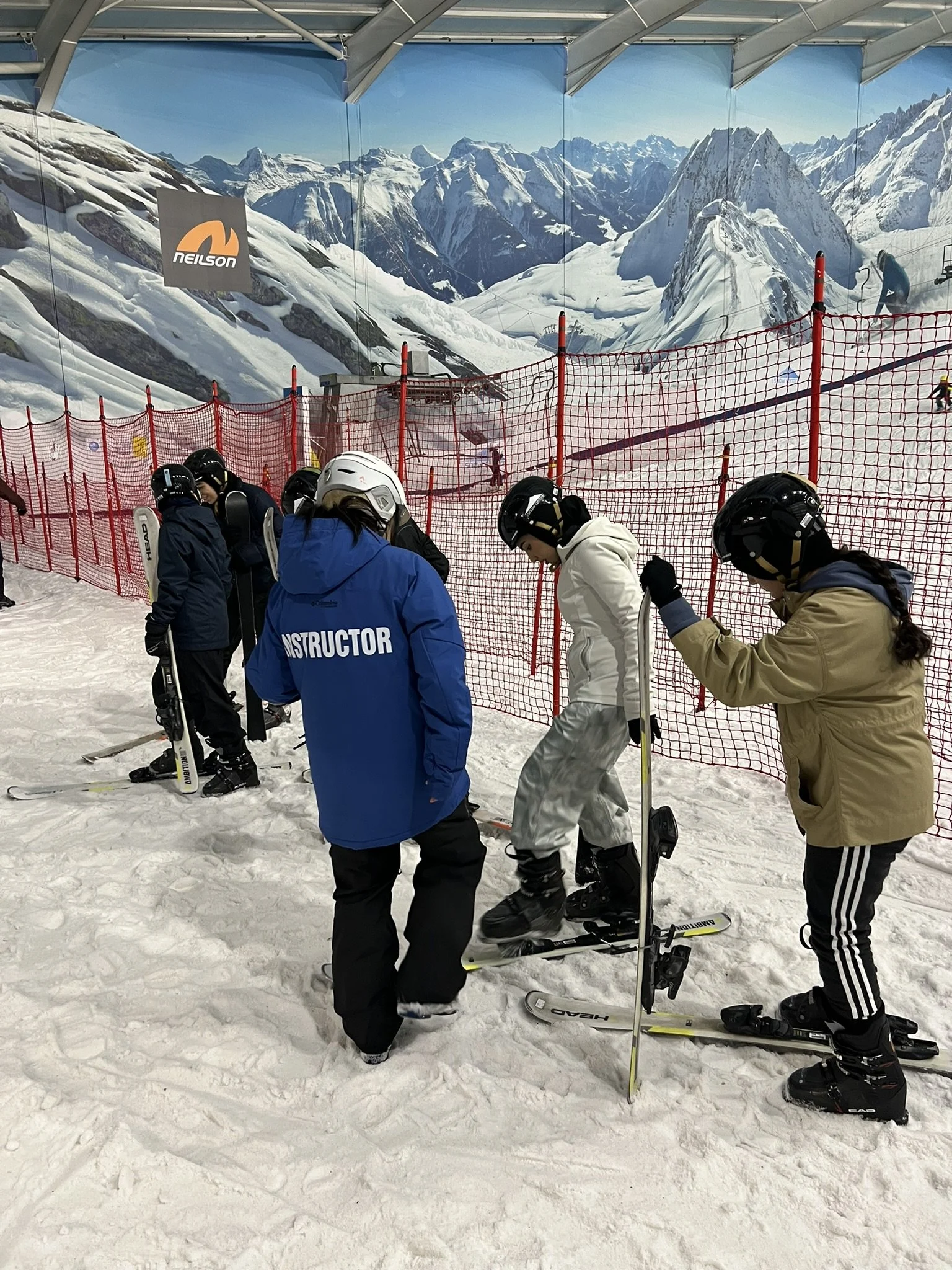 Guests at an indoor ski slope with mountain scenery mural, some wearing ski gear, instructor in blue jacket, helping a skier, with red safety netting