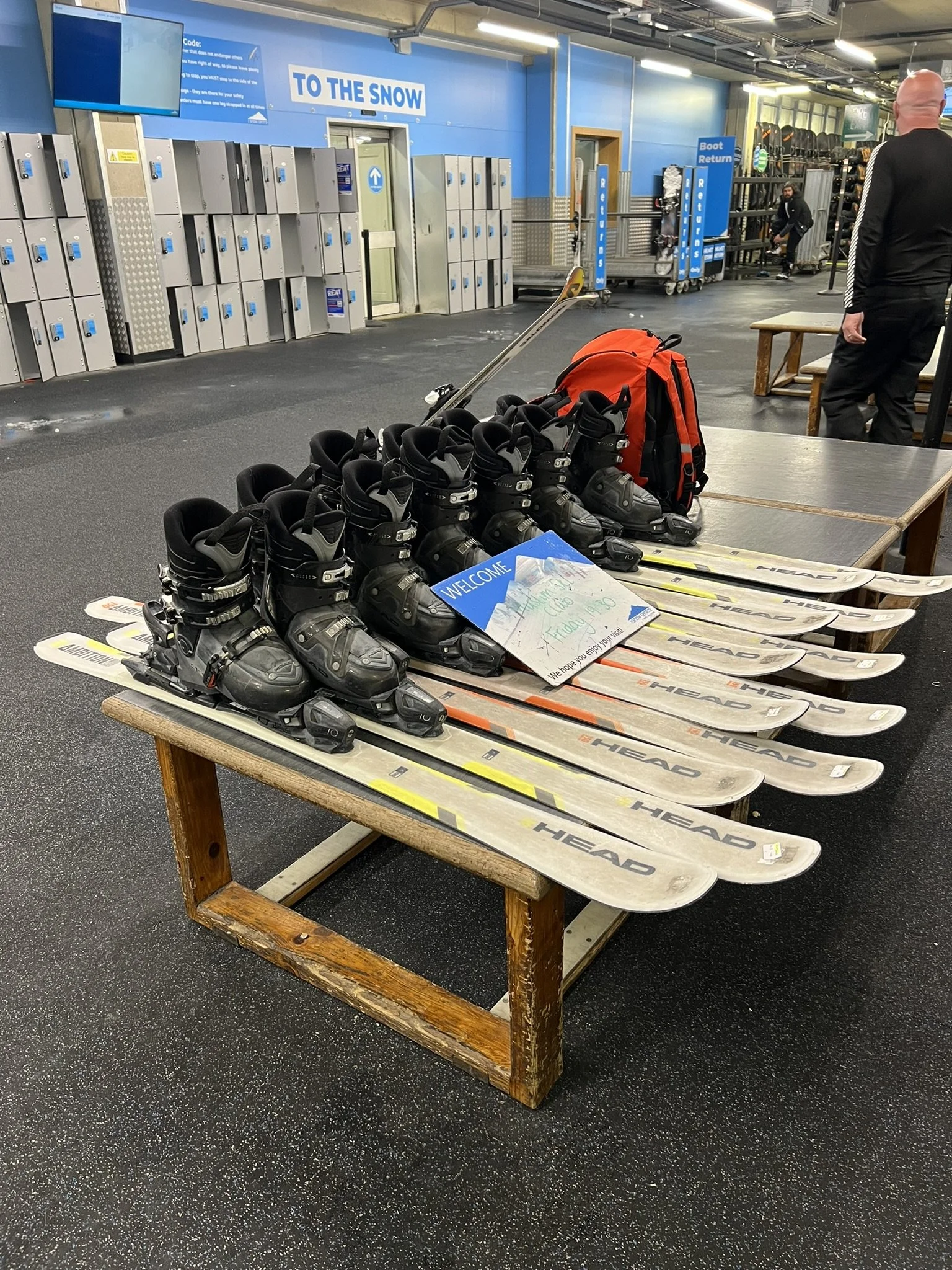 Ski boots, skis, and a backpack on a wooden table inside a ski rental shop, with lockers and signs in the background.