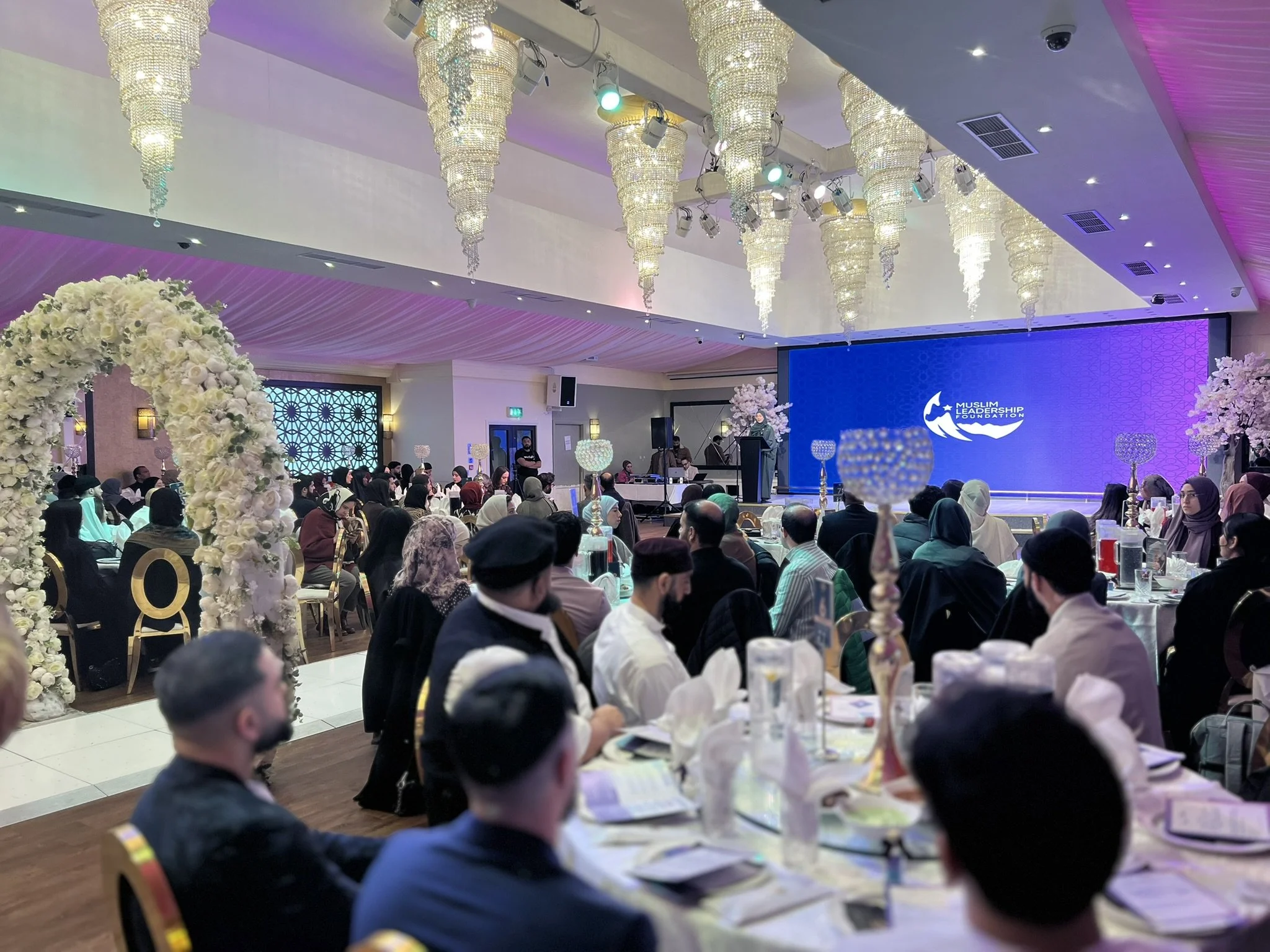 A large indoor event with many attendees seated at decorated tables, a stage with a large blue screen displaying the Muslim Leadership Foundation logo, floral arrangements, chandeliers, and stage lighting.