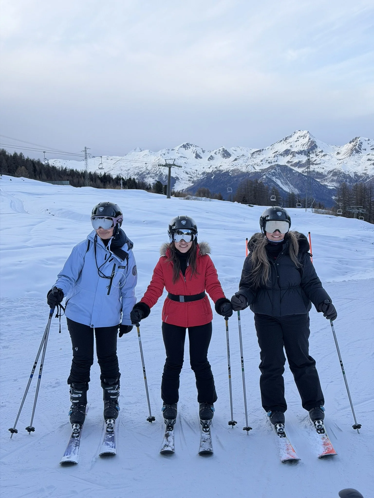 Three people in ski gear standing on a snowy mountain with ski lifts and snow-capped mountain peaks in the background.