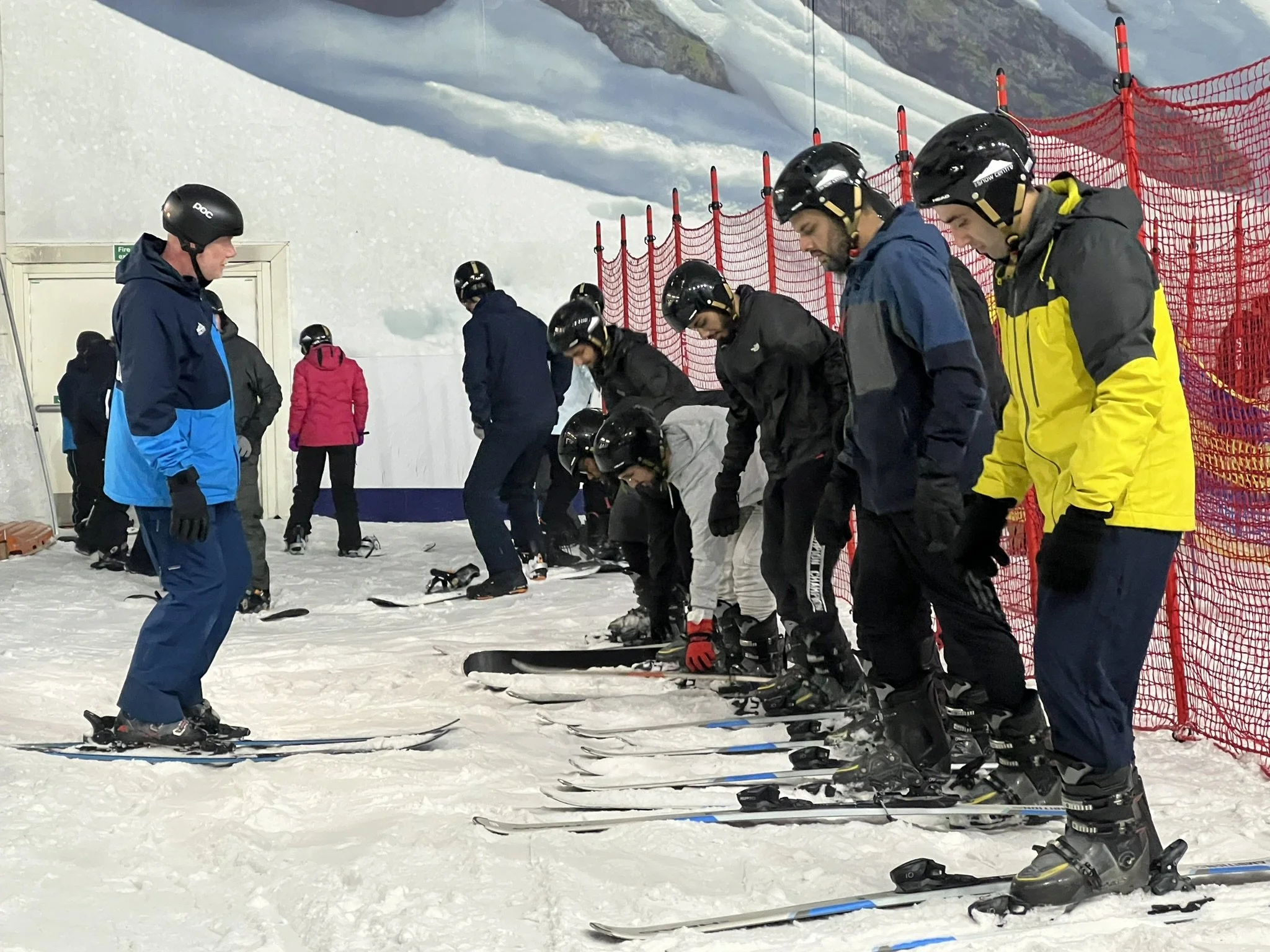 Group of skiers wearing helmets, standing in a line on an indoor ski slope with some adjusting their skis, with a ski instructor standing in front of them.