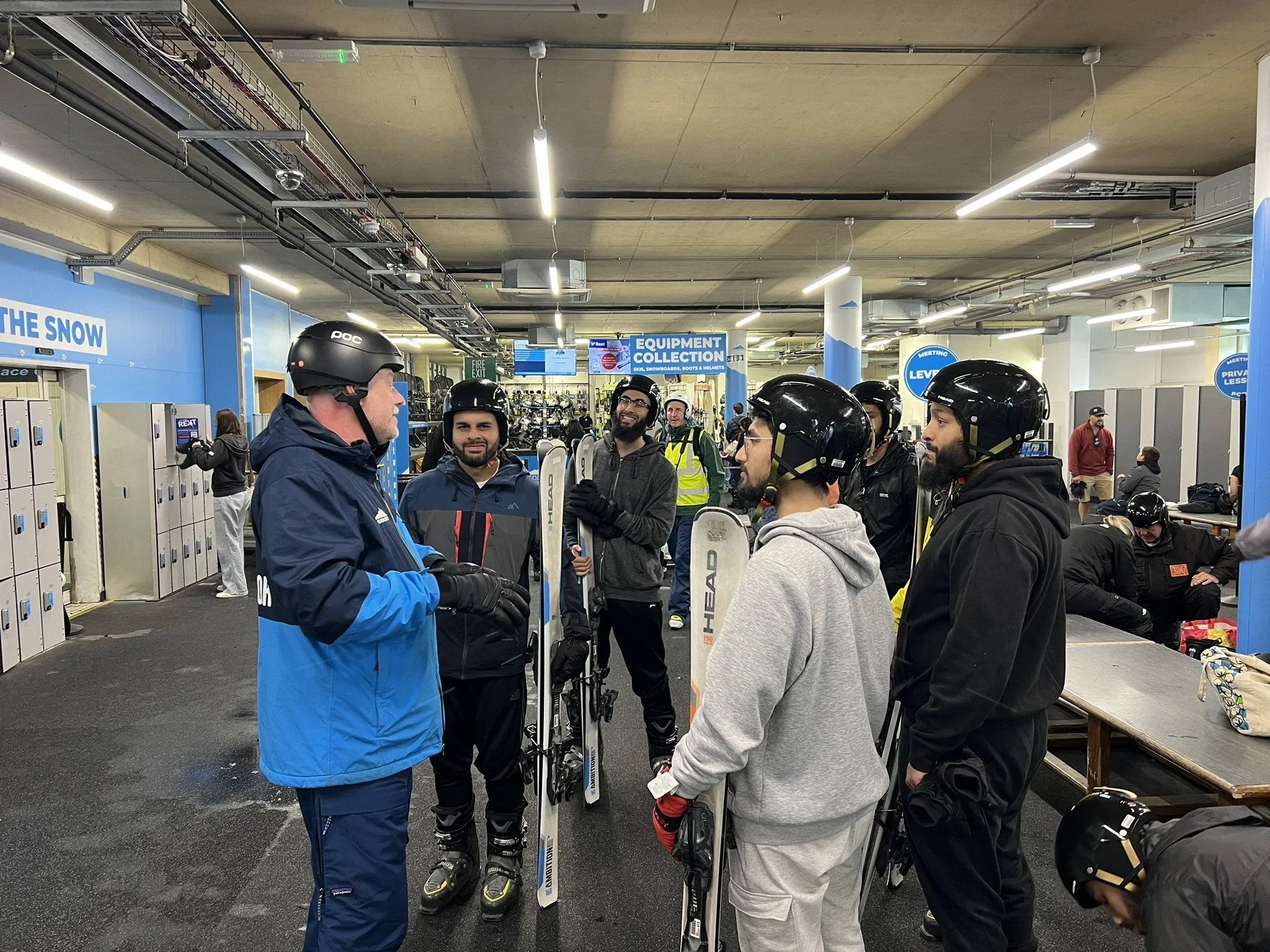 Ski instructor talking to a group of six skiers inside a ski rental shop, all wearing helmets, with lockers and ski equipment in the background.