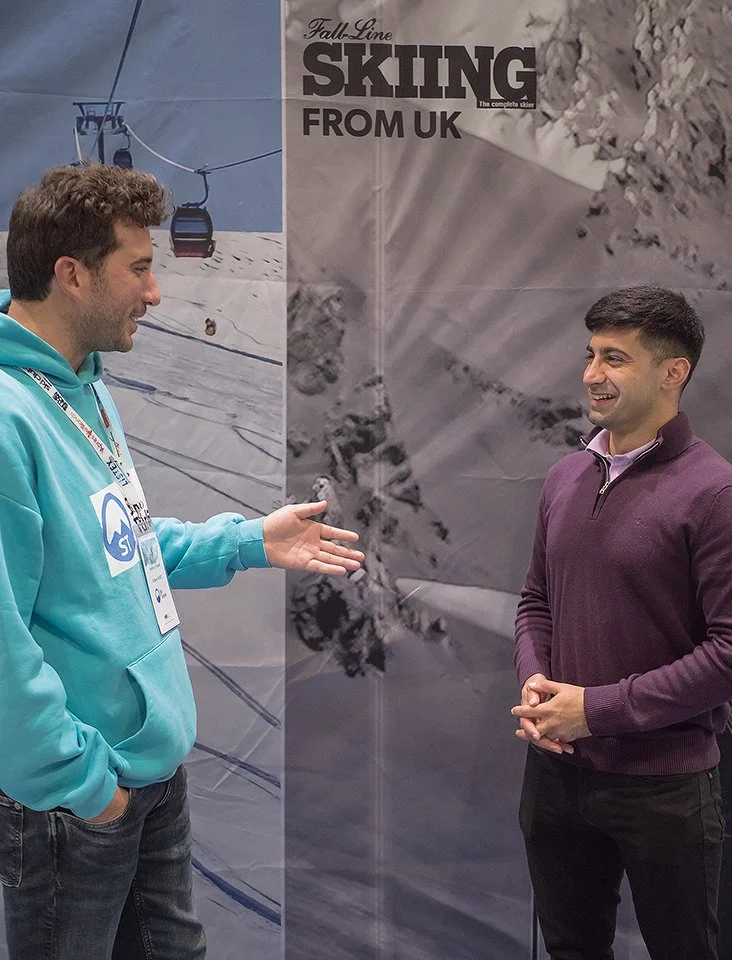 Two men are having a conversation indoors in front of a large poster advertising skiing from the UK. One man is wearing a turquoise hoodie with a ski event badge, and the other man is wearing a purple quarter-zip sweater. They are smiling and engagin